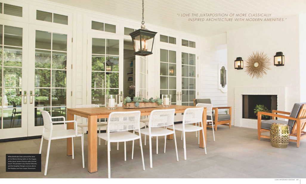 Bright modern dining room with white walls, large windows, a wooden dining table, and white chairs. Decor includes a black lantern hanging above, a fireplace with a small potted plant, and wall-mounted lanterns and a sunburst mirror.