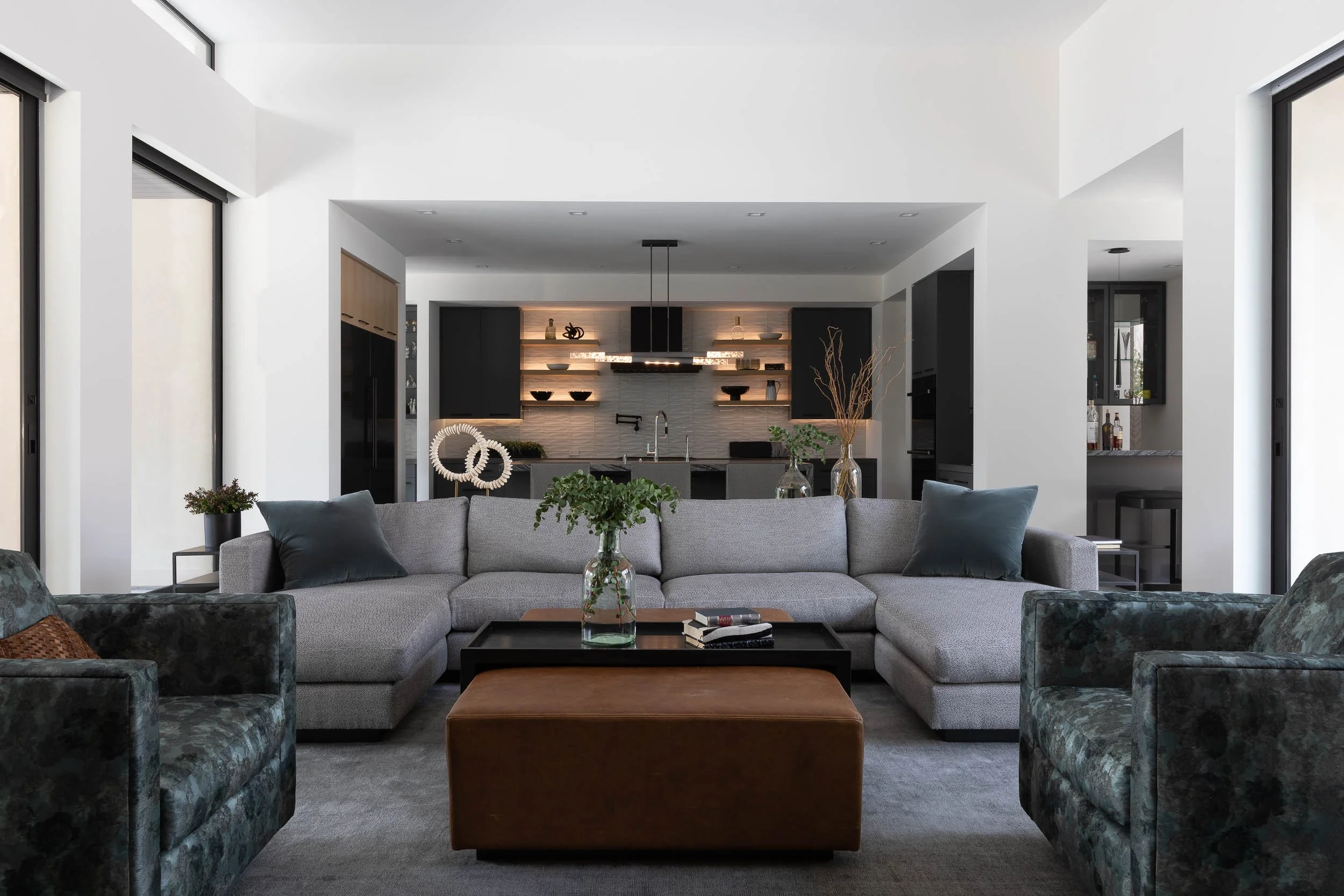 Modern living room with gray sectional sofa, glass vases with greenery, and a coffee table, open to a kitchen with black cabinets, open shelving, and pendant lighting, surrounded by large windows.