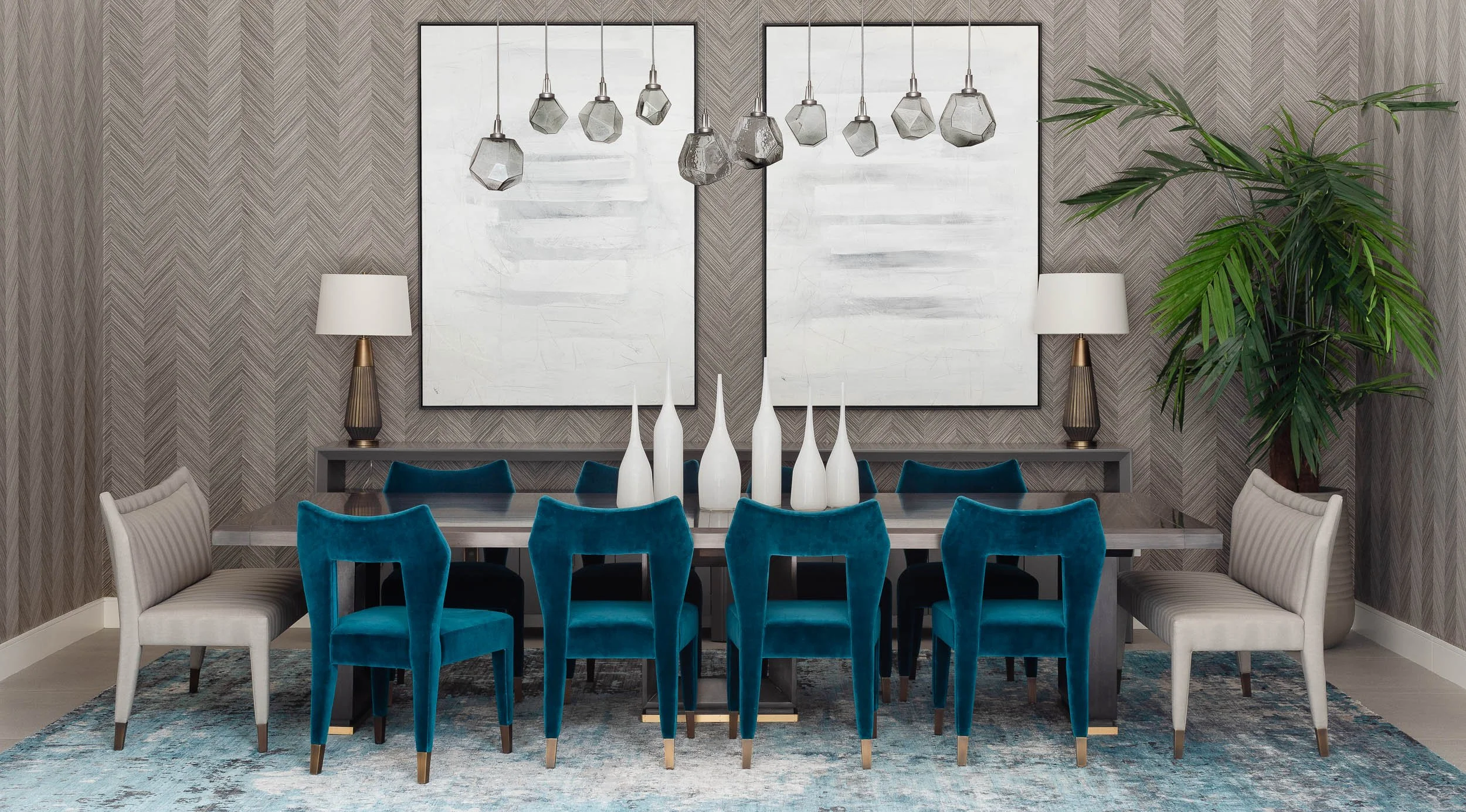 A modern dining room with a long table, eight blue velvet chairs, four white upholstered chairs, white vases centerpiece, and large framed abstract art on a textured gray wall, with two table lamps and a large green plant in the corner.