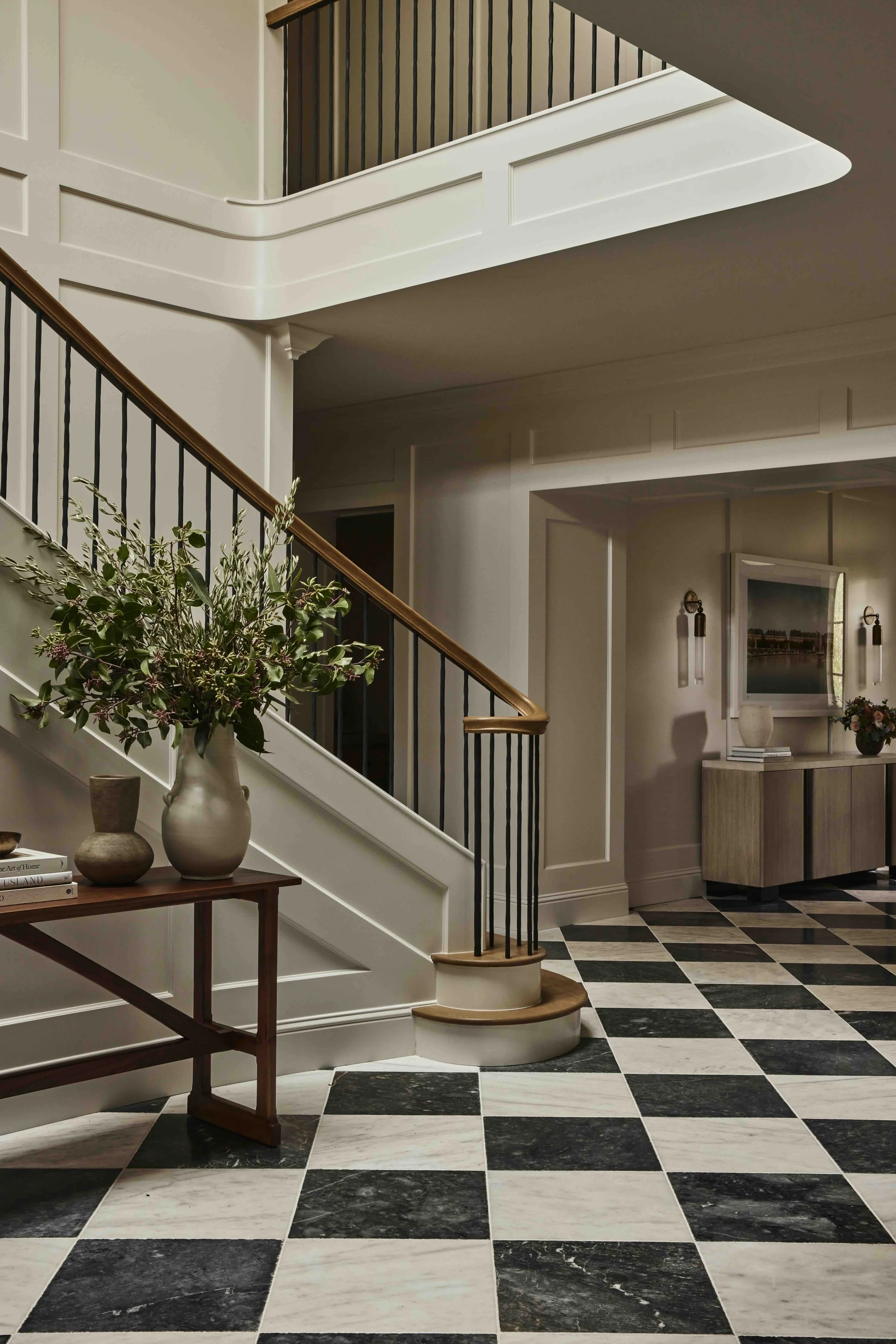 Interior of a house with black and white checkered marble flooring, a staircase with a wooden handrail, a console table with a large floral arrangement, and a wall-mounted TV above a wooden cabinet.