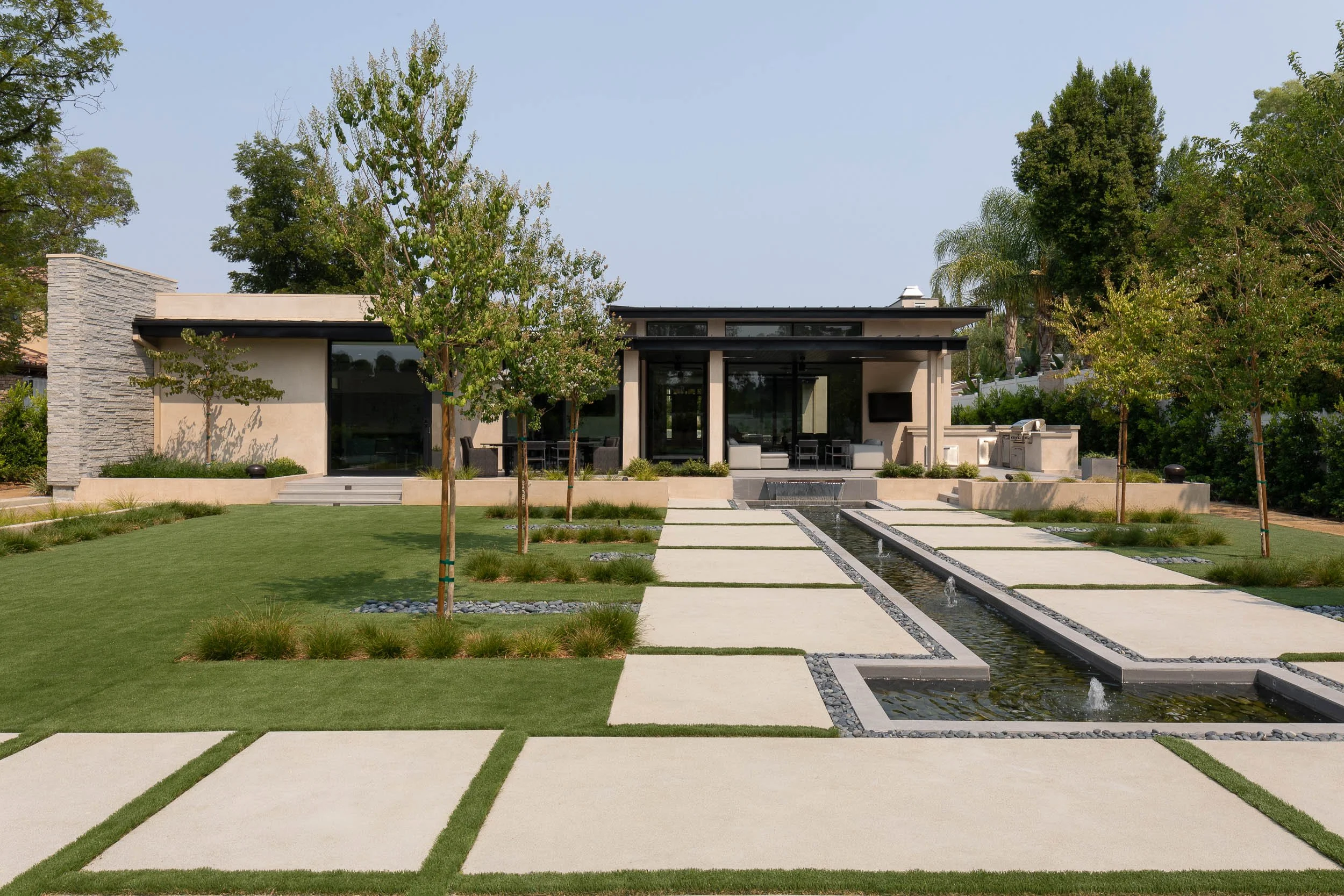 Modern house with a landscaped yard featuring a water feature, paved walkways, small trees, and outdoor seating area.
