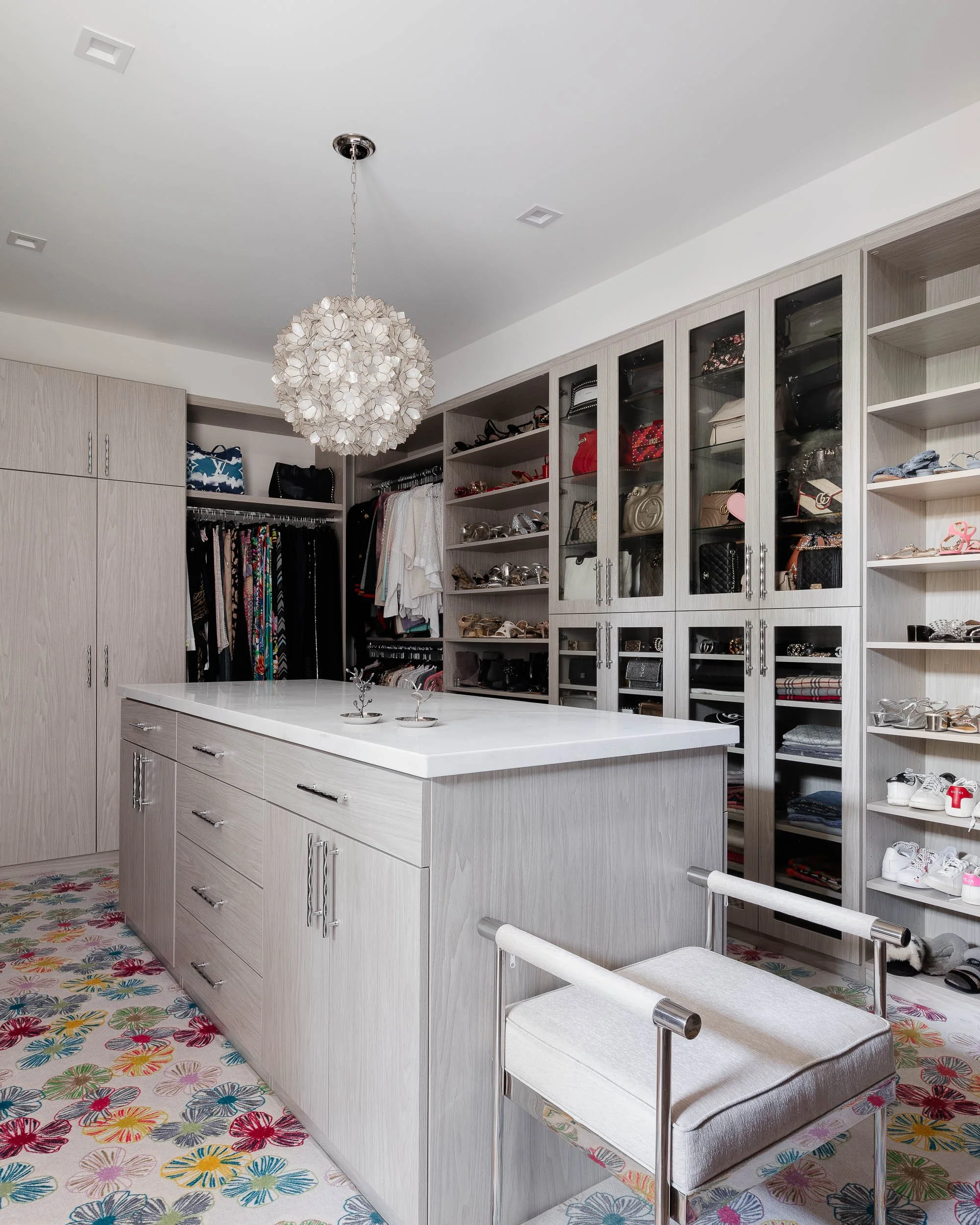 Modern walk-in closet with built-in shelves, glass-door cabinets, and a central island. Features clothing, handbags, shoes, and accessories. Includes a colorful floral rug and a white cushioned stool.