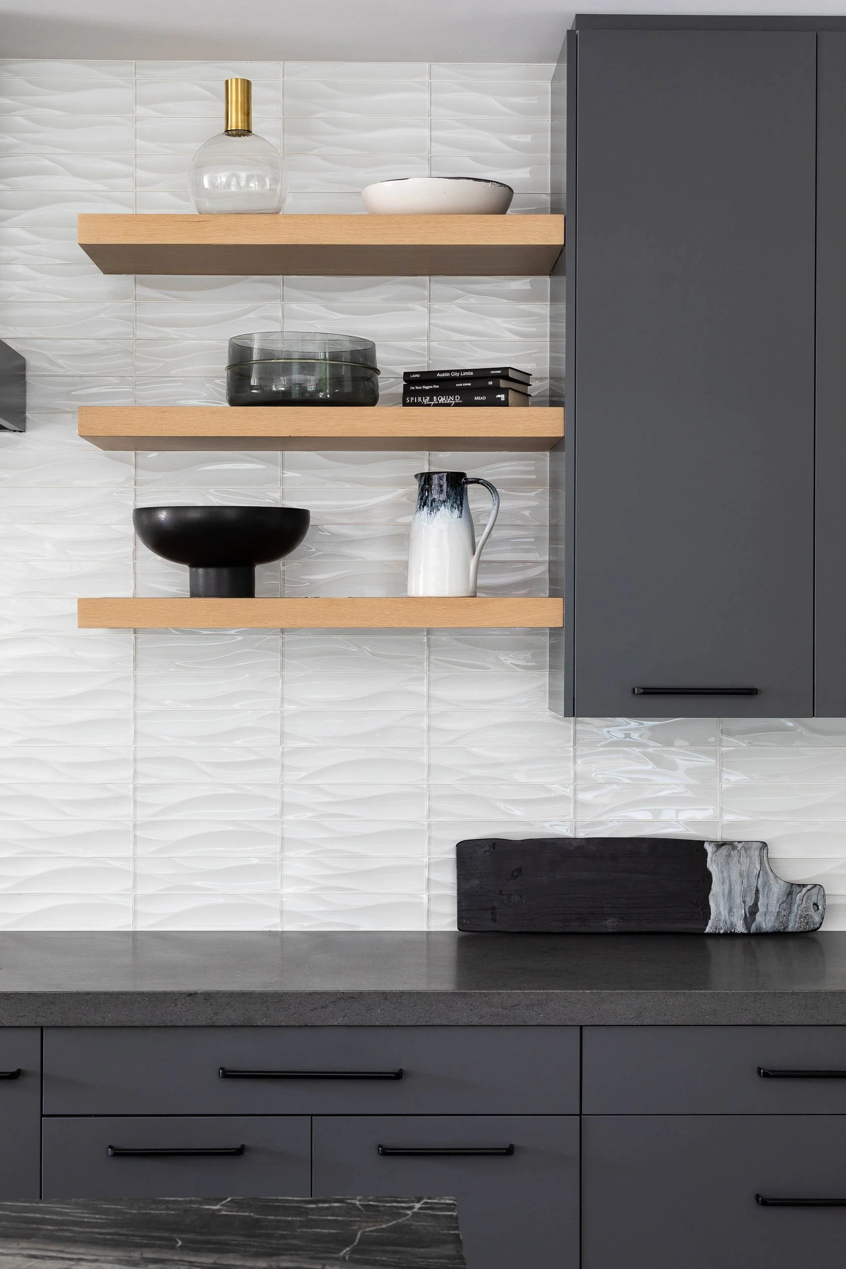 Modern kitchen with white textured tile backsplash, dark gray cabinets, and three minimalist wooden open shelves holding decorative items like vases, bowls, and books; a dark gray countertop with a black and white stone slab.