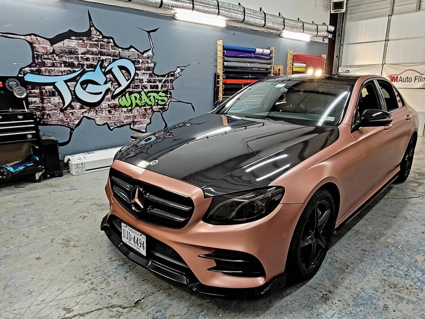 A Mercedes-Benz sedan with a matte pink body and black accents parked inside an auto detailing garage. The garage has a blue wall with graffiti art, shelves with rolls of wrap film, and some tools and equipment nearby.