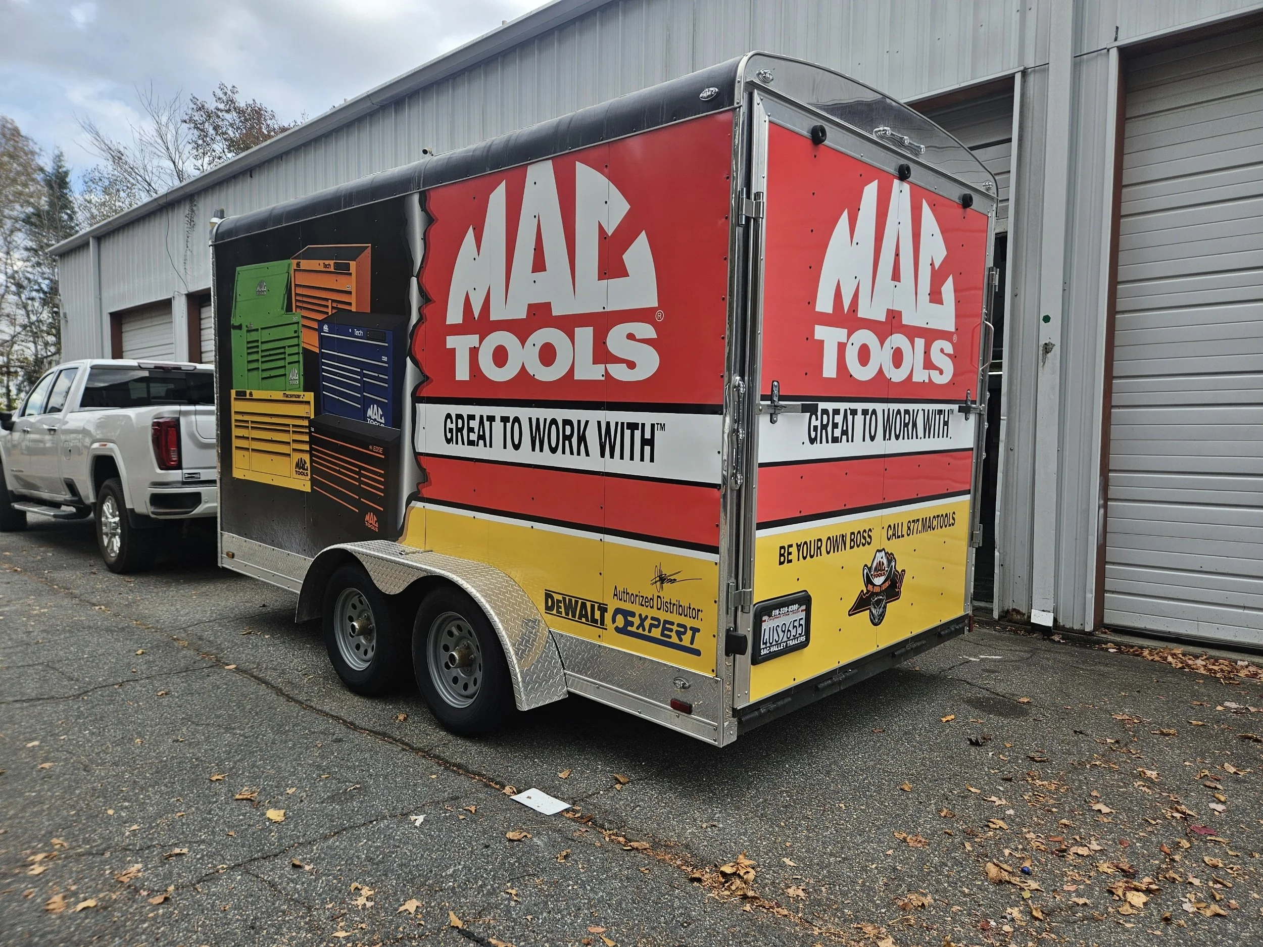 A trailer advertising MAC Tools with colorful images of tool chests and logos, parked next to a building with a white pickup truck nearby.