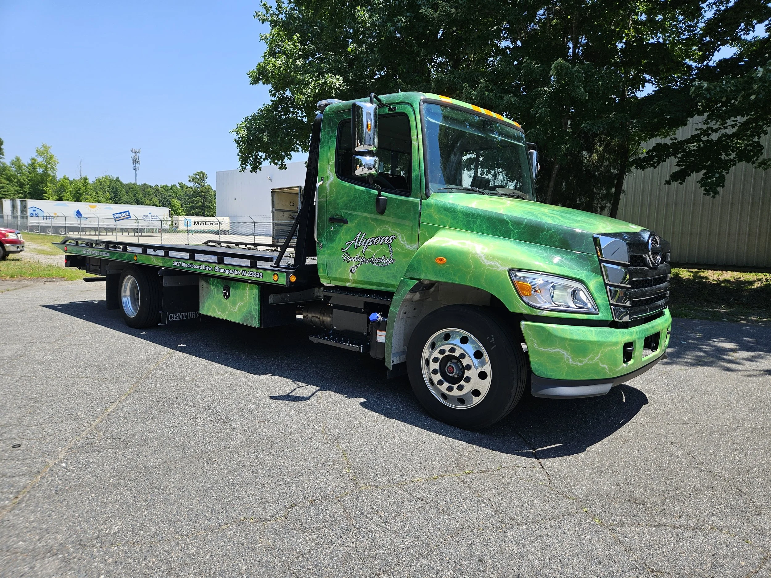 Green flatbed tow truck parked on asphalt with trees and a fence in the background.