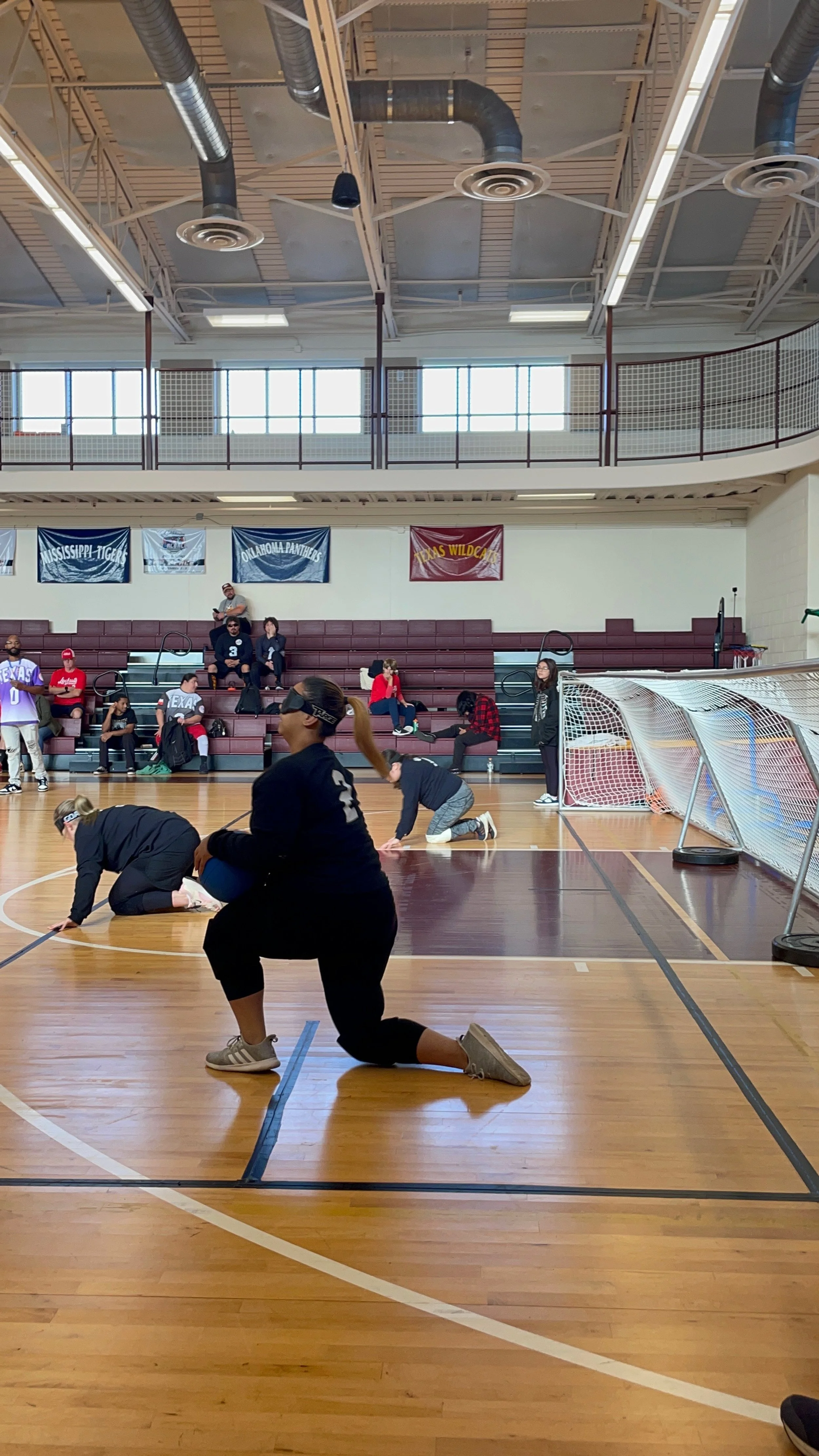 People participating in a goalball game inside a gymnasium, with some kneeling and others sitting on bleachers watching.