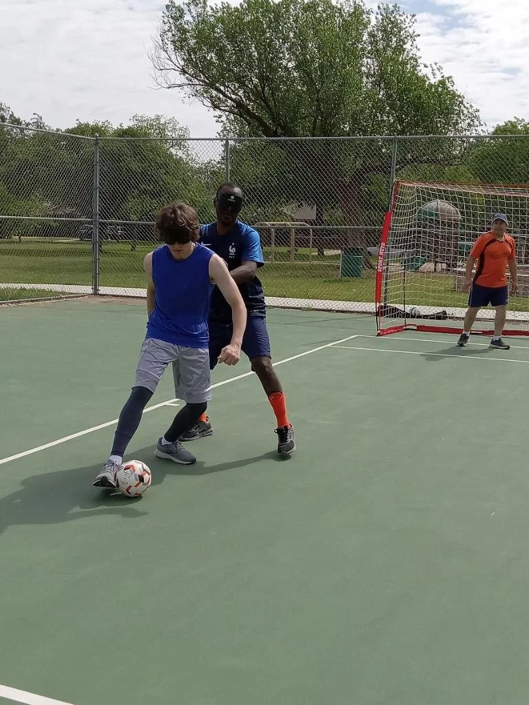 Two boys playing soccer on a tennis court, one in a blue shirt and gray shorts with a soccer ball, the other in a dark blue shirt and shorts defending, with a third person in an orange shirt and shorts watching near a soccer goal.