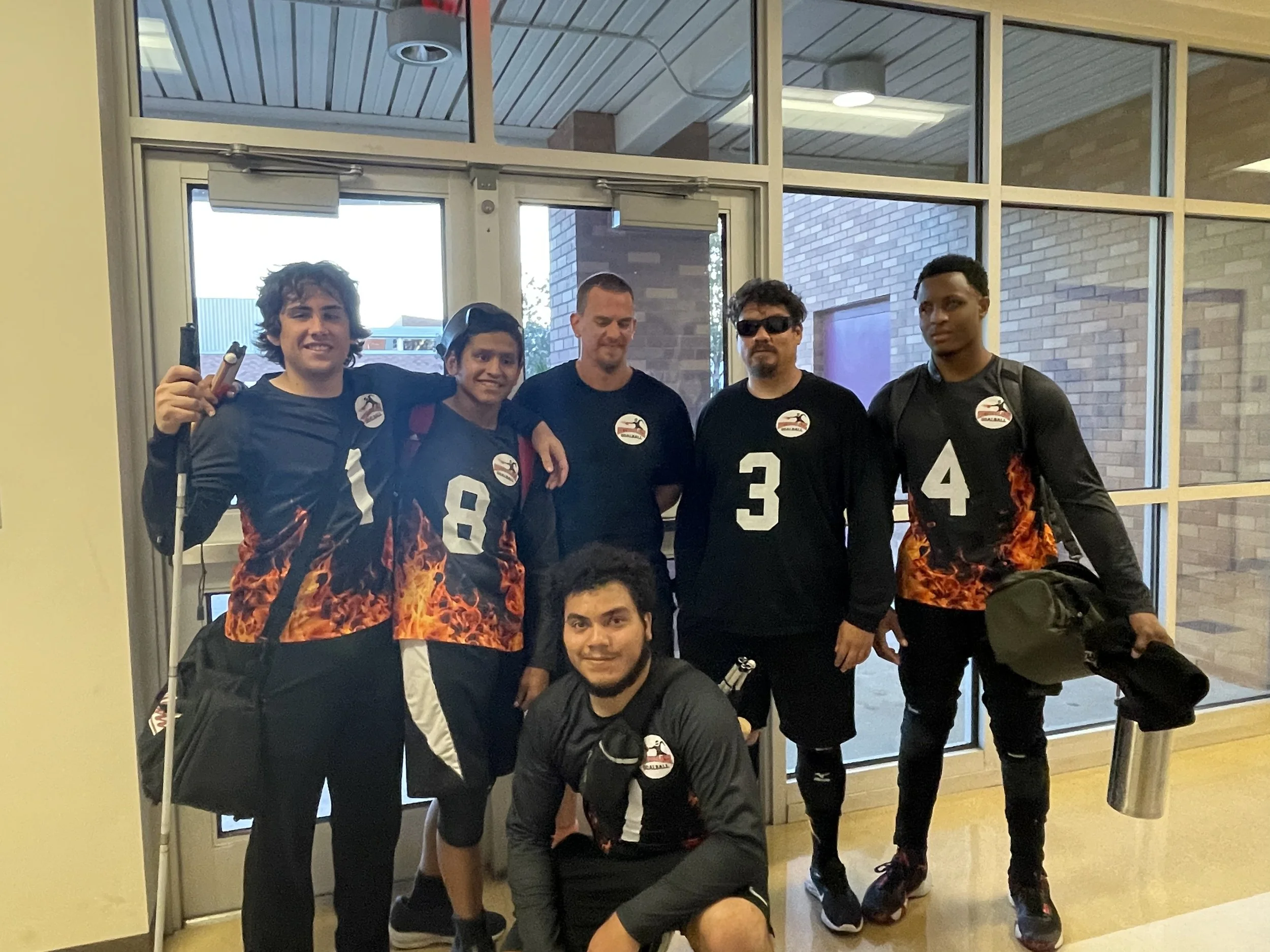 Group of six people, five standing and one kneeling, in a sports hall, wearing sports jerseys with flame designs, holding sports equipment, posing for a photo.