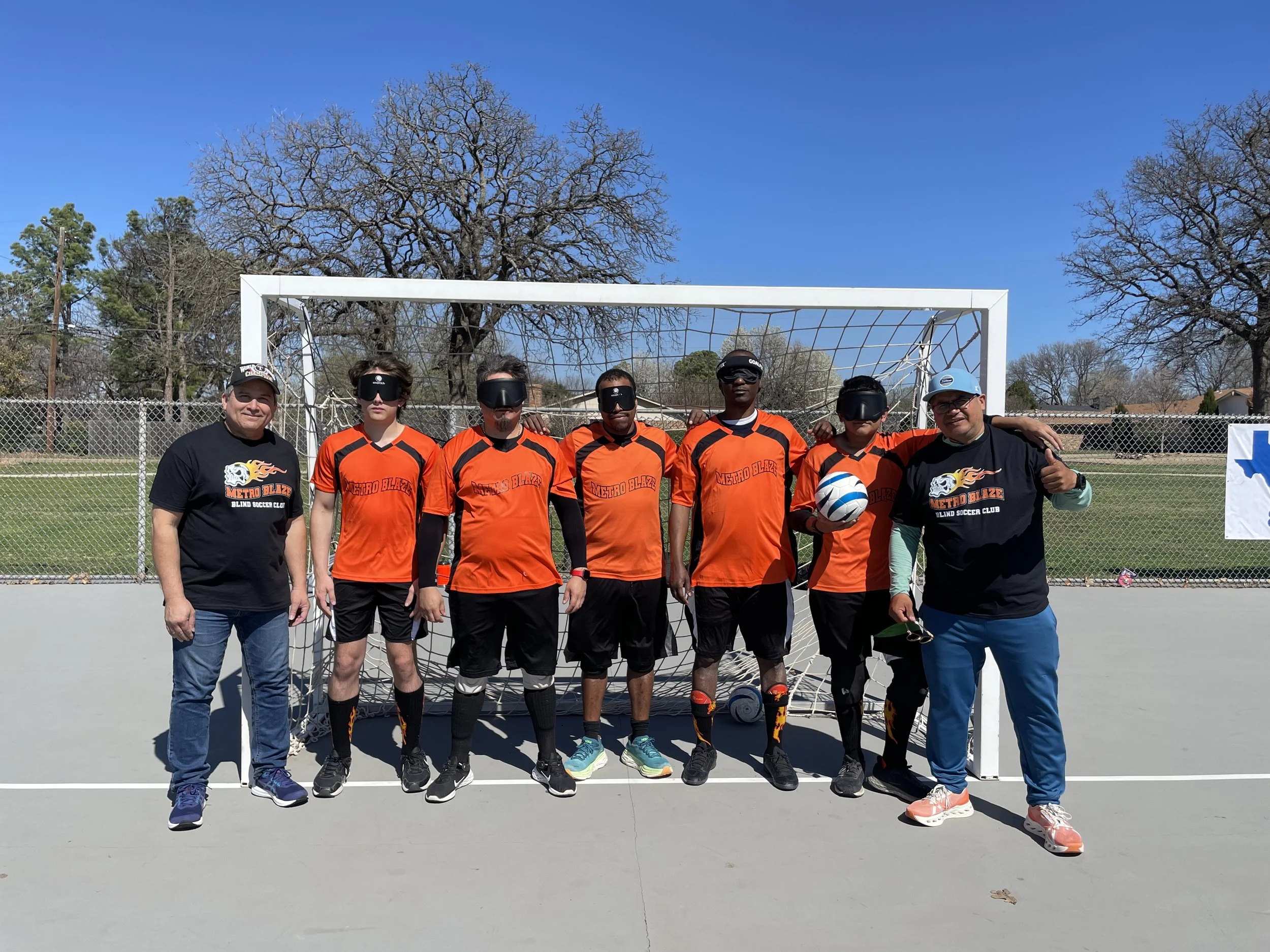 Group of seven people, five soccer players in orange uniforms and two coaches, standing in front of a soccer goal on an outdoor field, with trees and blue sky in the background.