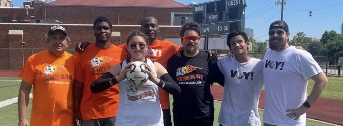 Group of six people standing on a soccer field, with one woman holding a soccer ball. They are smiling and posing for a photo, some wearing matching orange and white shirts that say 'Metro Blue' and 'V.O.Y!'