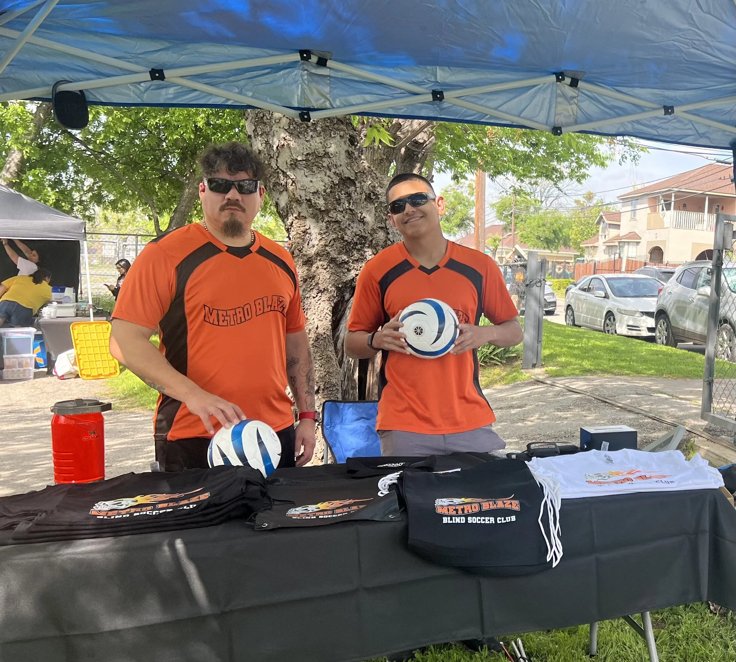 Two men in orange and black sports jerseys standing behind a table at an outdoor event, holding soccer balls, under a blue canopy with a tree in the background.