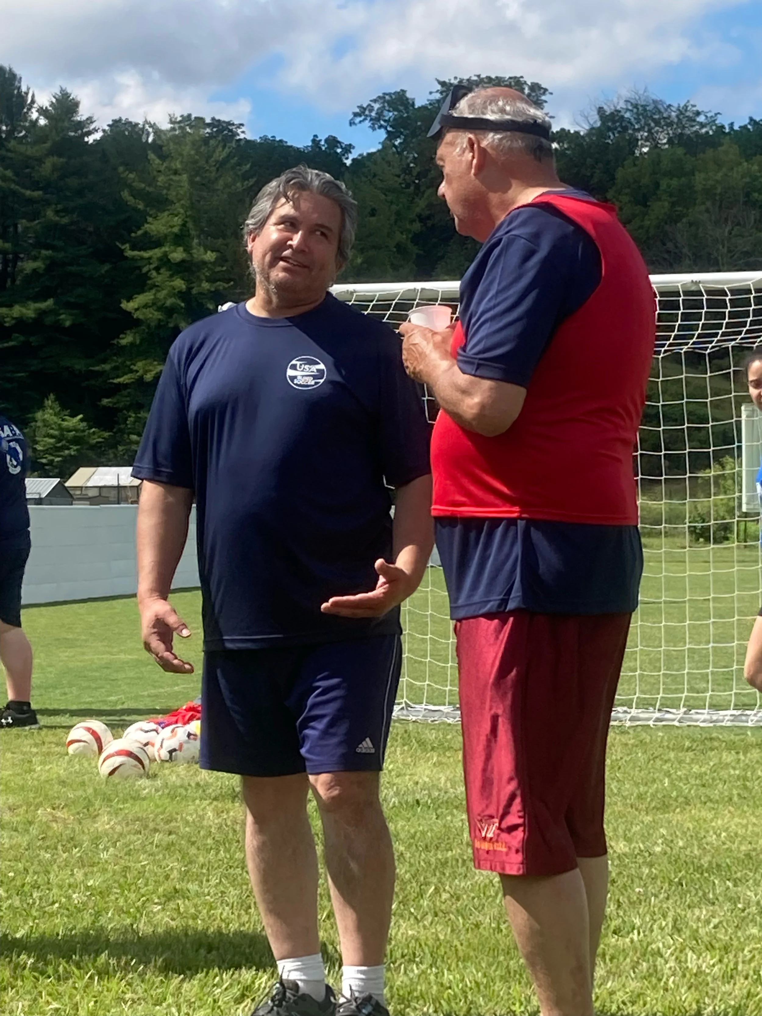Two men having a conversation on a soccer field, with a goal net in the background and several soccer balls on the ground. One man is wearing a navy blue athletic shirt and shorts, and the other is in a red shirt and matching shorts, holding a cup.