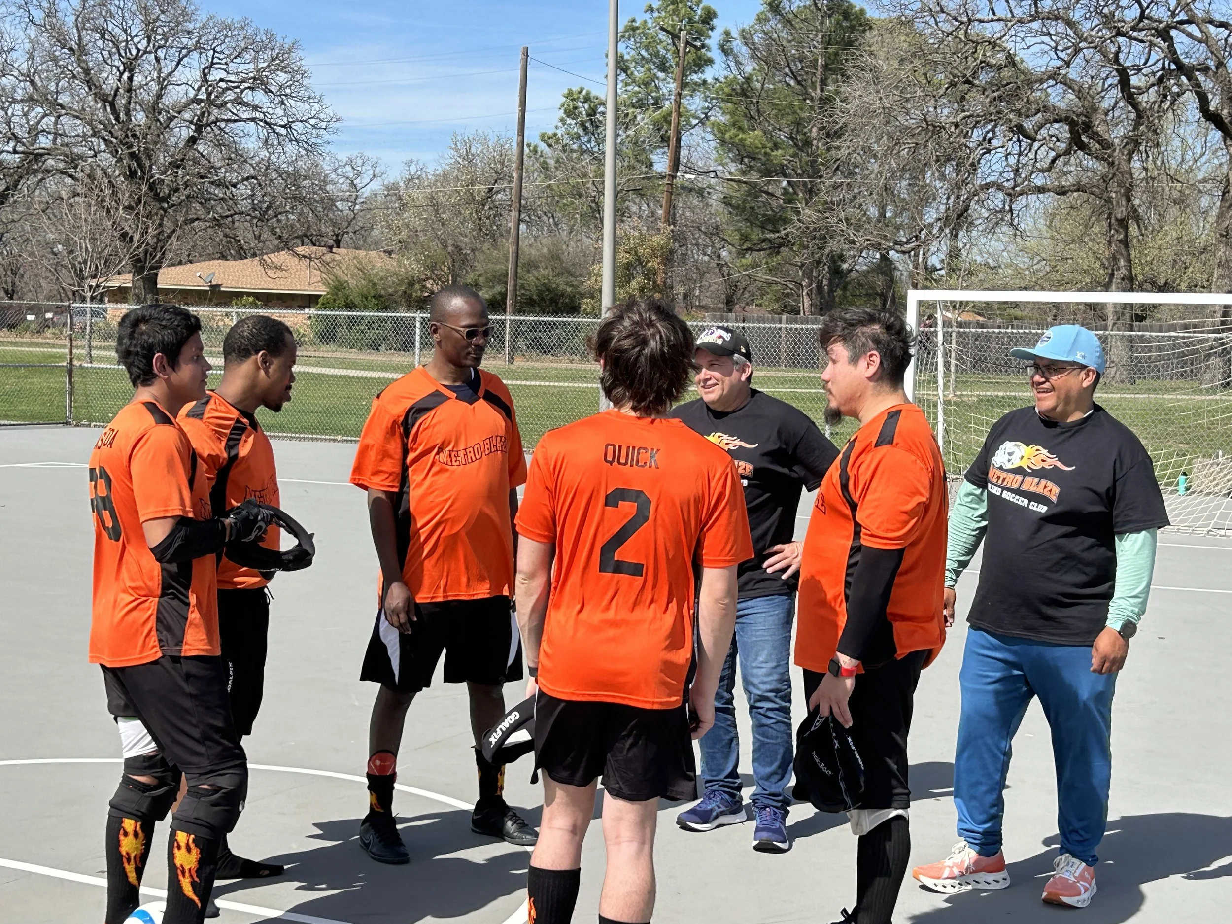 A group of people gathered on an outdoor sports court, some wearing orange sports jerseys and others in black shirts, while talking before a soccer game.