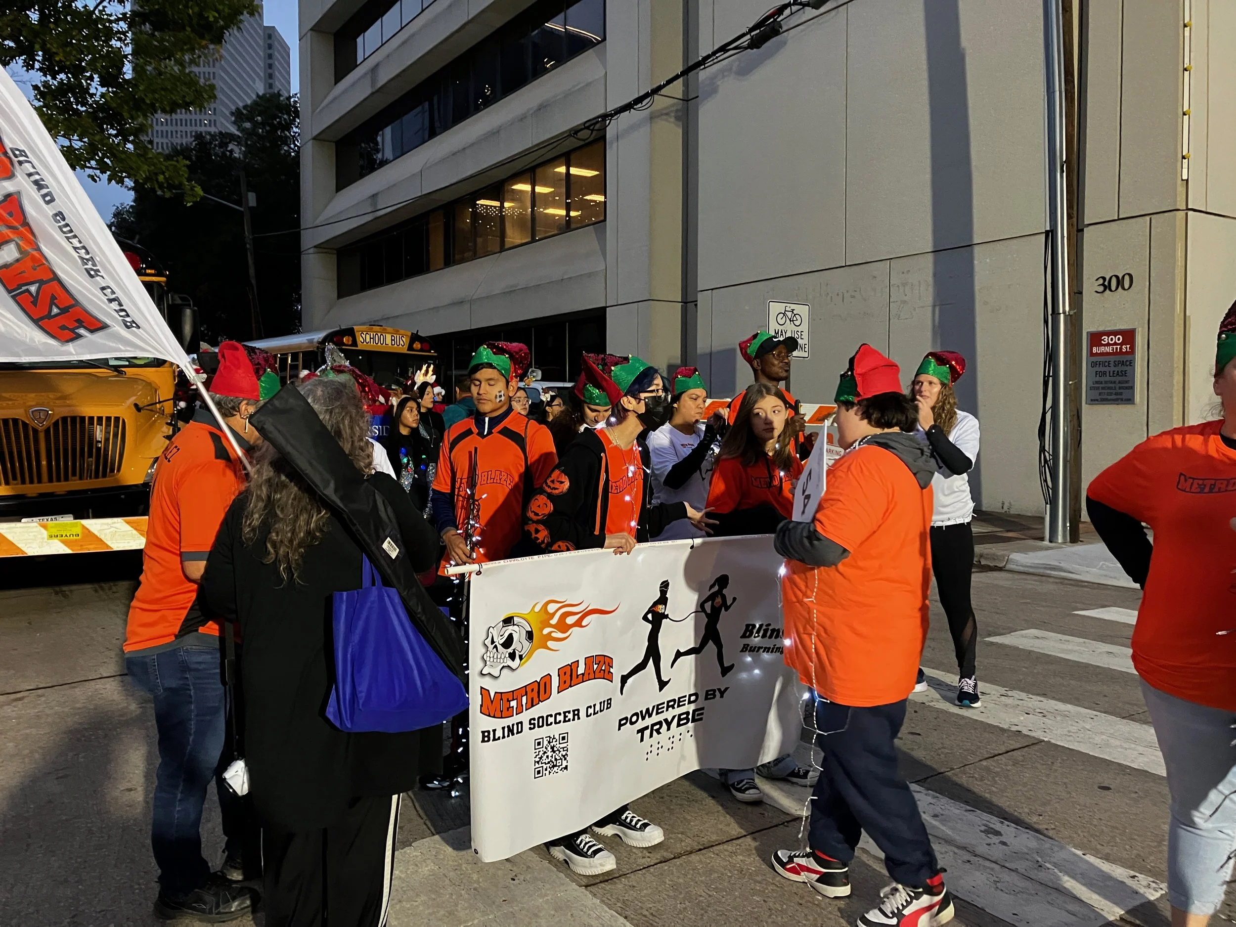 Metro Blaze and Blind Fury Runners wearing elf hats and orange jackets holding a banner in a city street during evening. A school bus is parked nearby, and there are tall buildings in the background during the Fort Worth Parade of Lights 2023