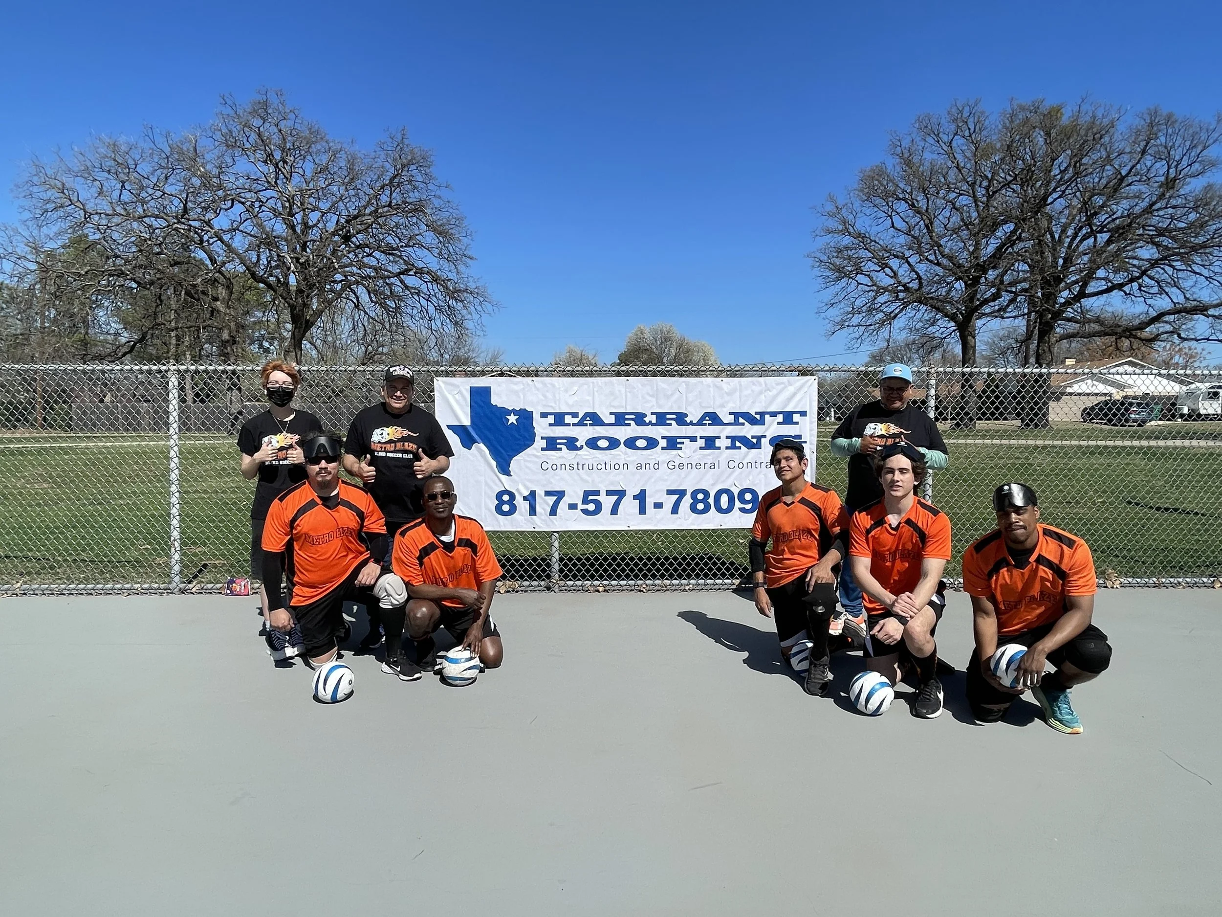 A group of seven people, some kneeling and some standing, posing in front of a chain-link fence with a banner for Tarrant Roofing. The people are wearing sports jerseys and holding footballs, on an outdoor sports court with trees and houses in the ba