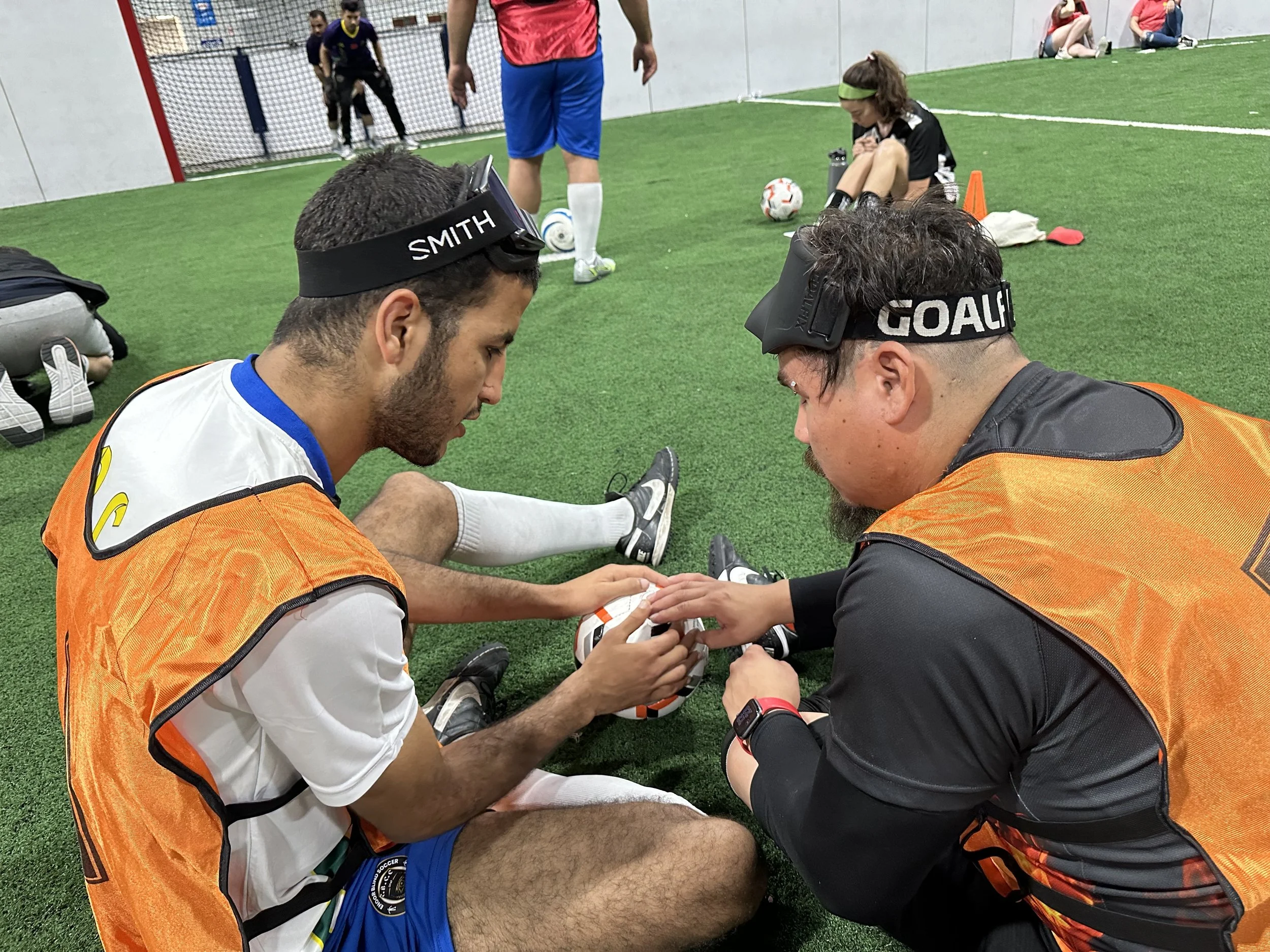 Two soccer players sitting on the ground, one wearing a white jersey and the other a black jersey with orange vests, are holding and inspecting a soccer ball. They are on an indoor soccer field with a green turf, surrounded by people, some sitting an