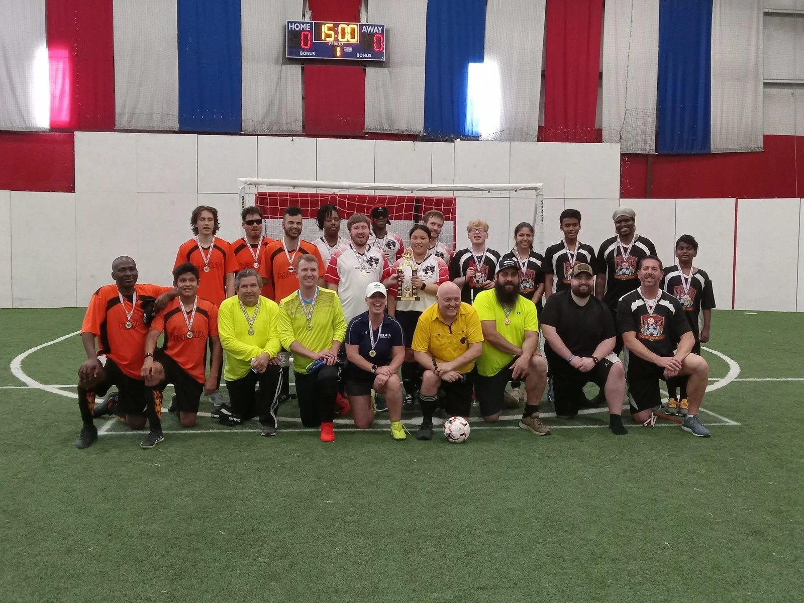 Group of people, including players and coaches, posing with medals and a trophy on an indoor soccer field, with a scoreboard showing 15 minutes remaining in the game.