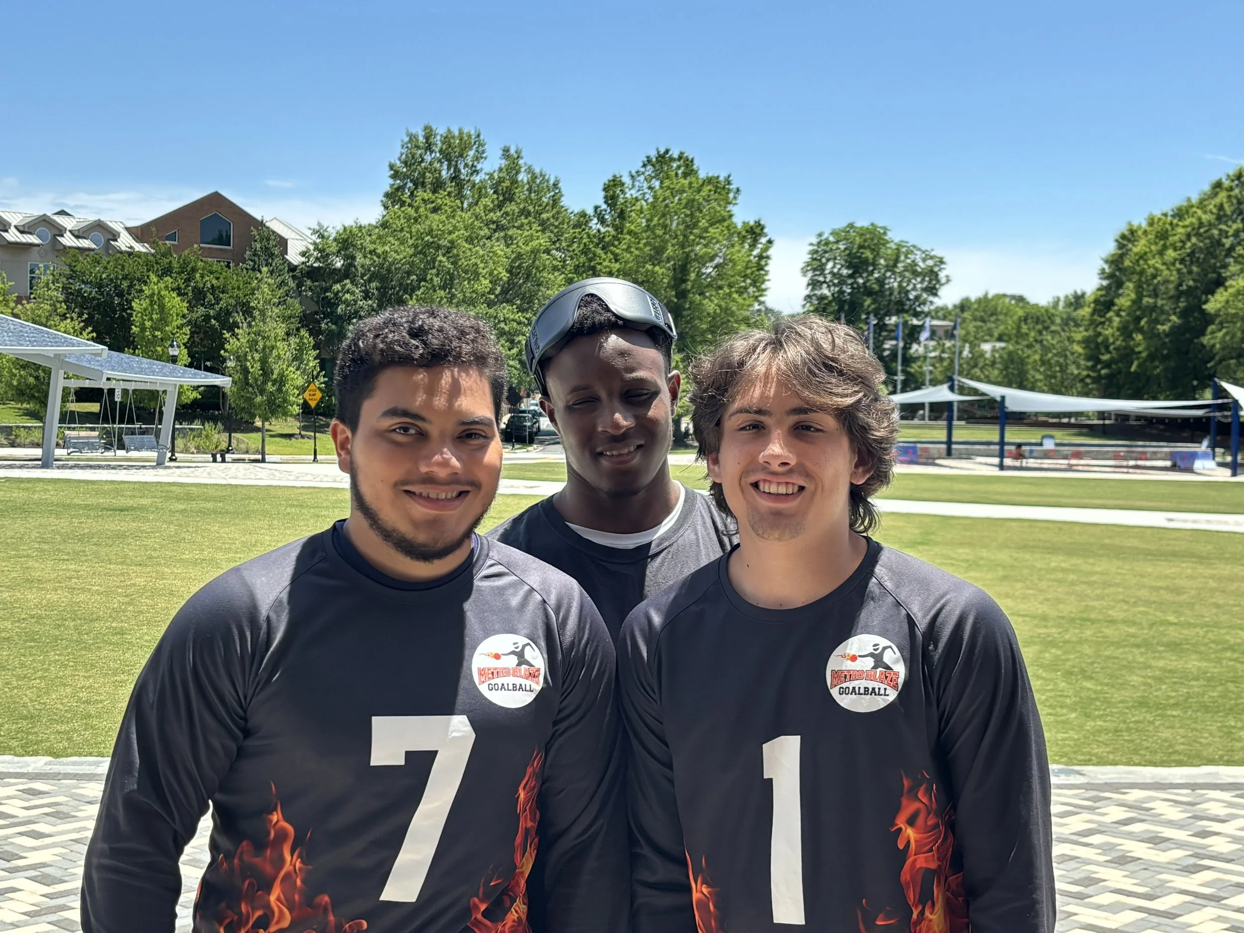 Three young men in goalball jerseys standing outdoors on a grassy area, smiling, with trees and a park in the background.