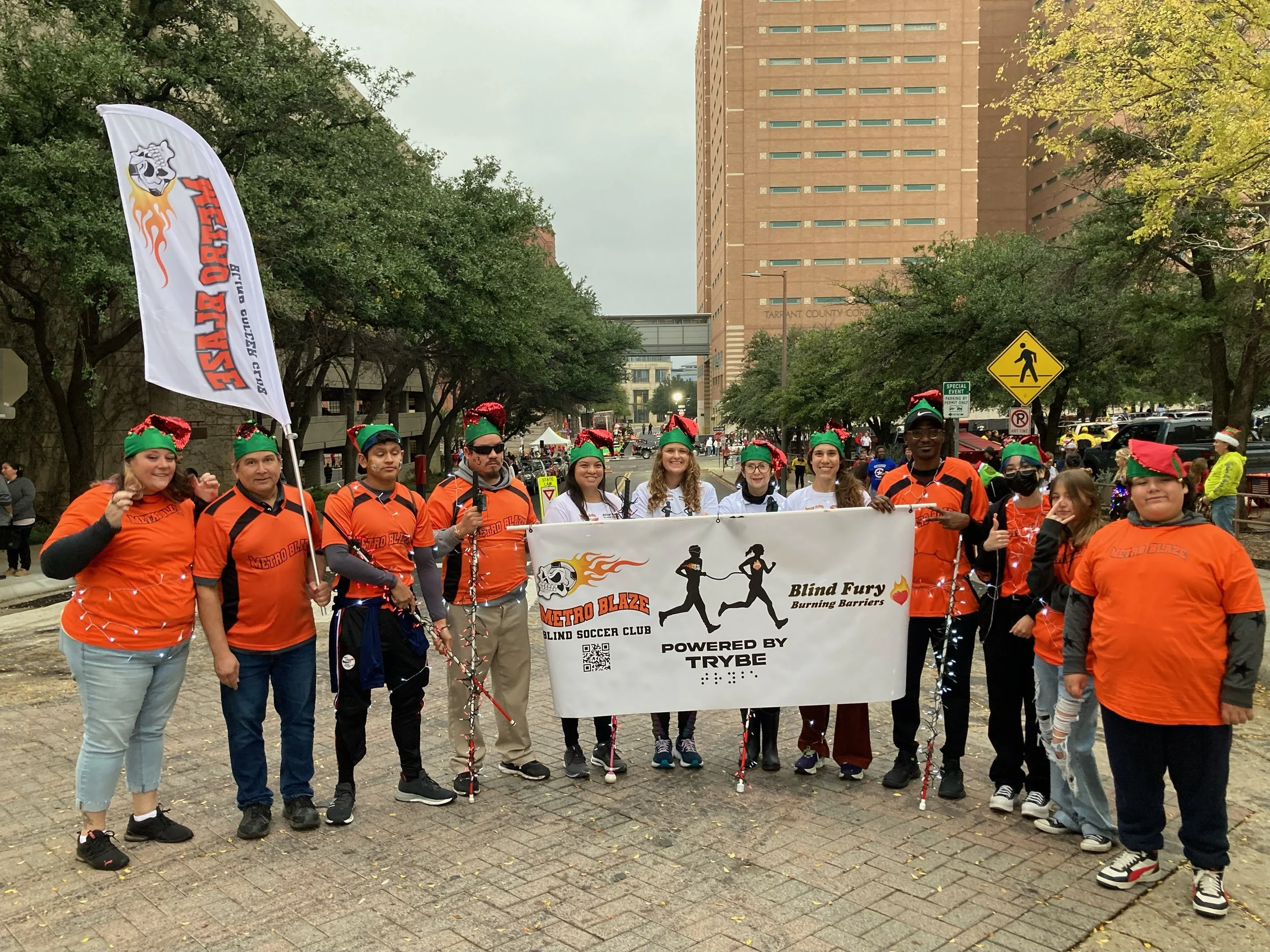 Group of people dressed in orange shirts and green Santa hats standing on a sidewalk holding a banner and a flag with a logo of a skull with flames, a running figure, and the words "Metro Blaze Blind Soccer Club." The scene appears to be during a par