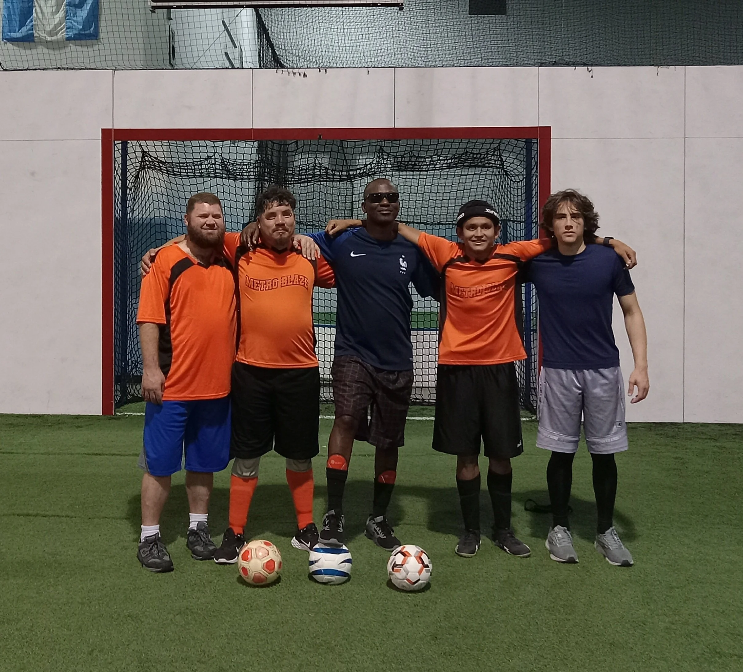 Group of five young men standing on an indoor turf soccer field, posing in front of a goal net with four soccer balls in front of them.