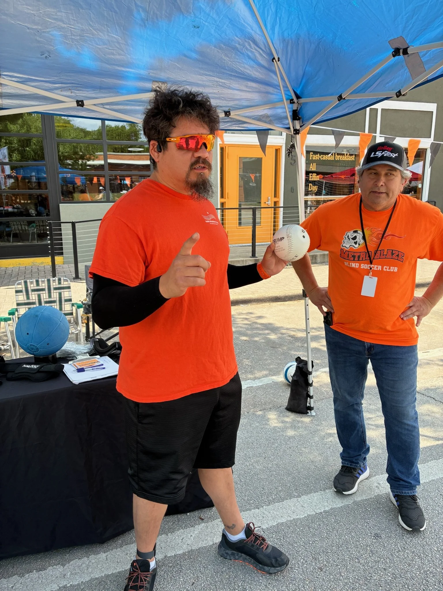 Two men standing outside under a blue canopy, one holding a baseball, both wearing orange shirts, with a table that has a blue ball and some papers, and a building with large windows behind them.