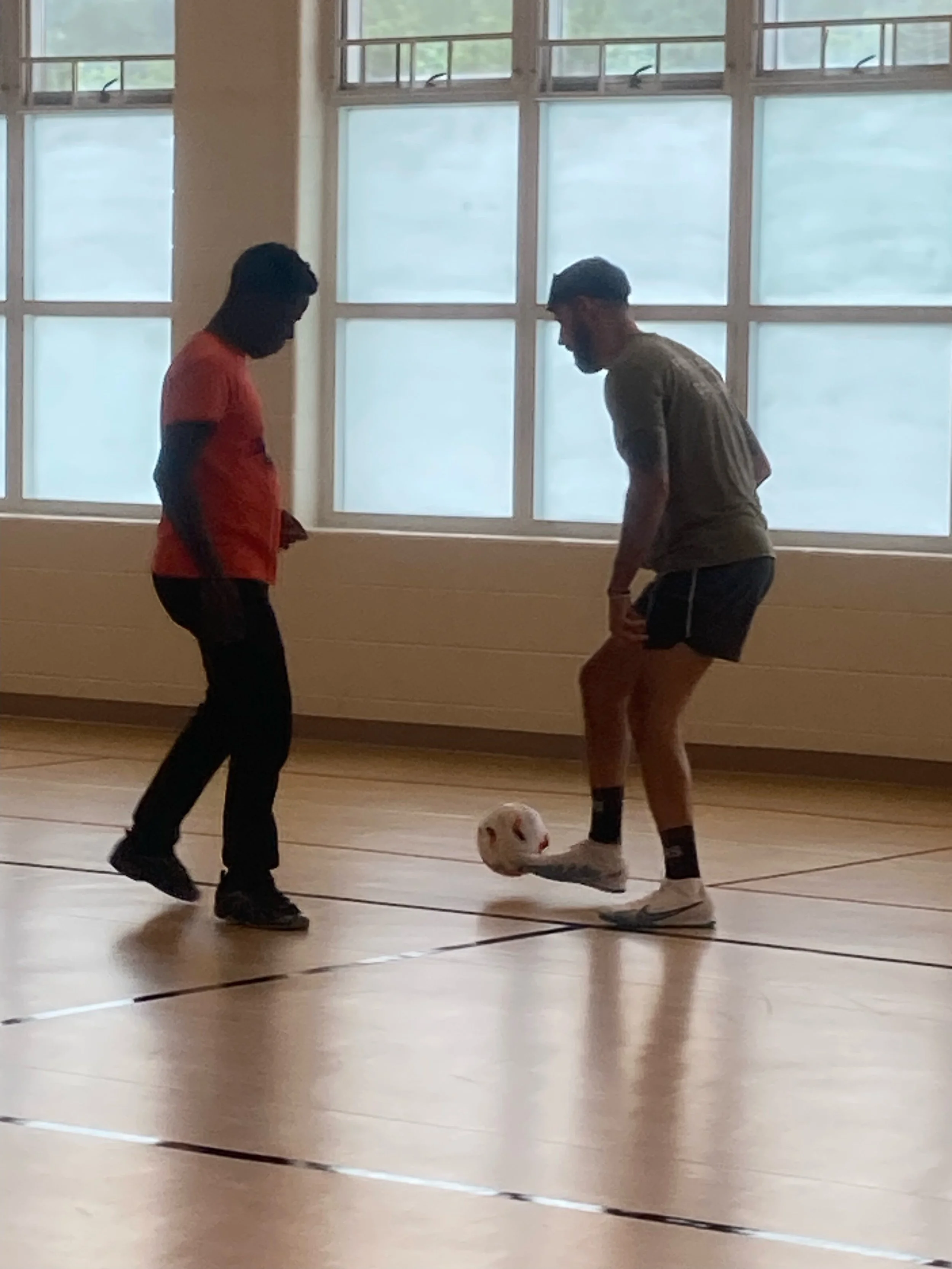 Two men playing indoor soccer with a ball, one is wearing a red shirt and dark pants, the other in a gray shirt and shorts, in a gymnasium with large windows.