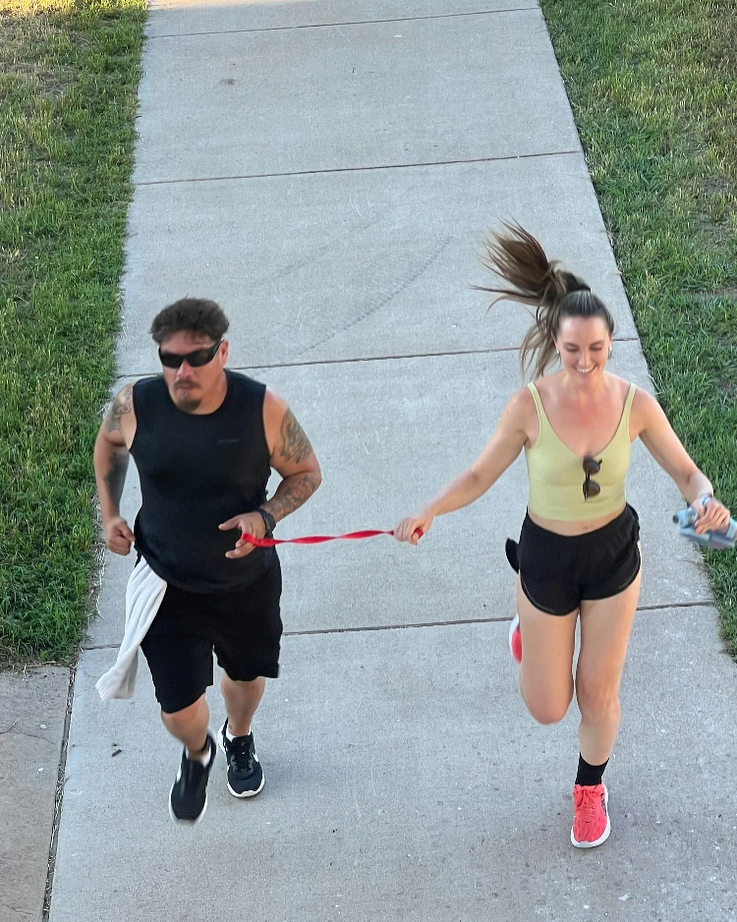 A man and a woman running on a sidewalk, holding a red tether between them, with the woman's hair flying in the air as she smiles.