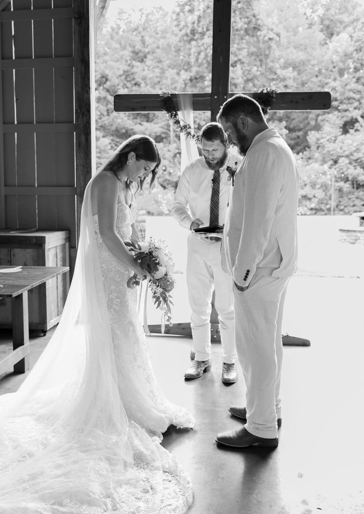 A black and white photo of a bride and groom during their wedding ceremony, standing indoors with an officiant reading from a tablet, near a large cross decorated with flowers, with trees visible outside.