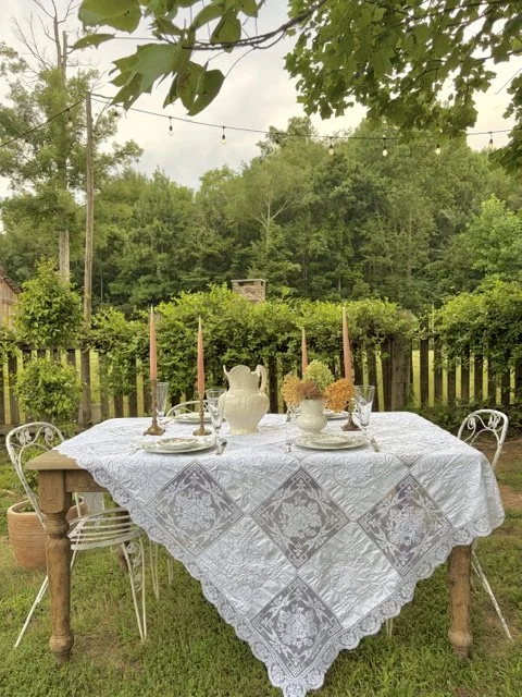 Outdoor dining table set with a white lace tablecloth, vases with flowers, candles, and glassware, surrounded by trees and a wooden fence, with string lights overhead.