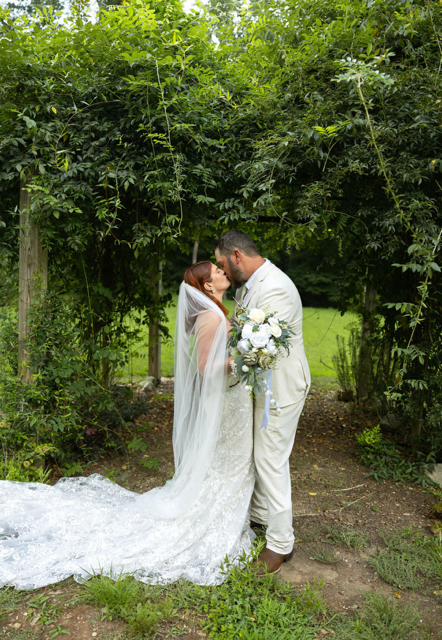 A bride and groom share a kiss outdoors beneath a green, leafy archway, with the bride holding a bouquet of white flowers.