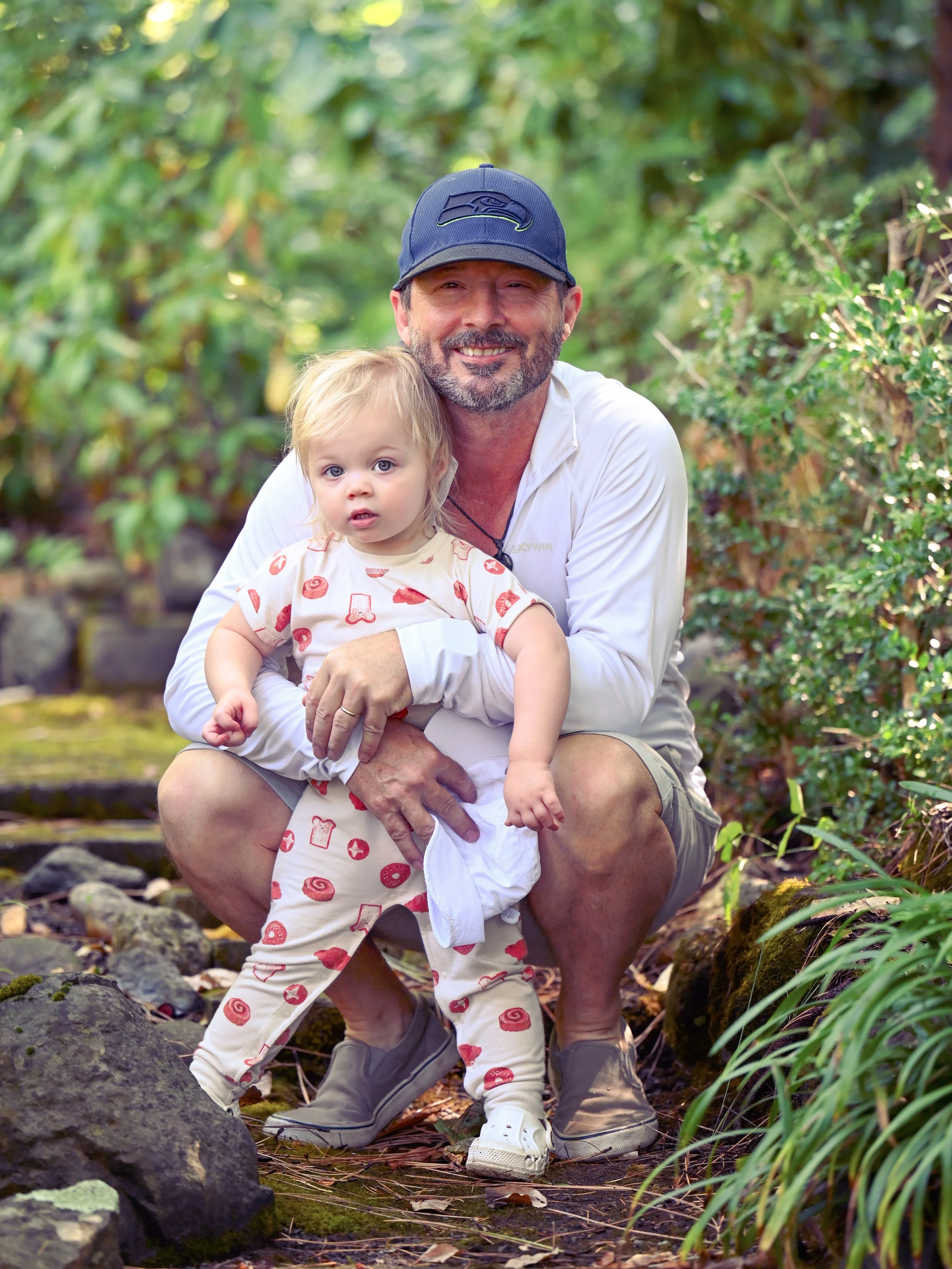 A man kneeling outdoors with a young girl, both looking at the camera, surrounded by green foliage.