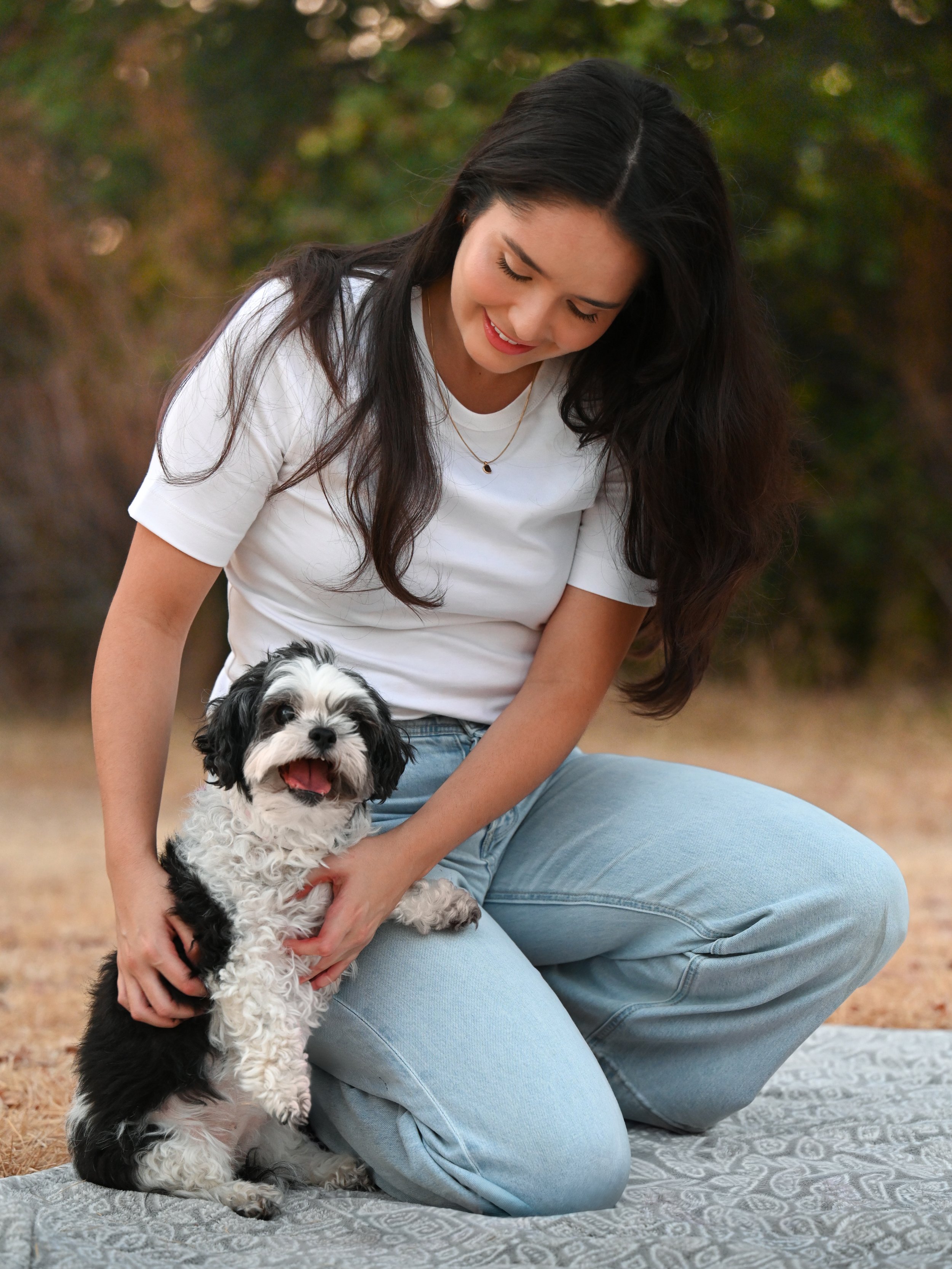 A woman with long dark hair wearing a white t-shirt and light blue jeans kneels on a blanket outdoors, smiling at a small black and white puppy that she holds gently.