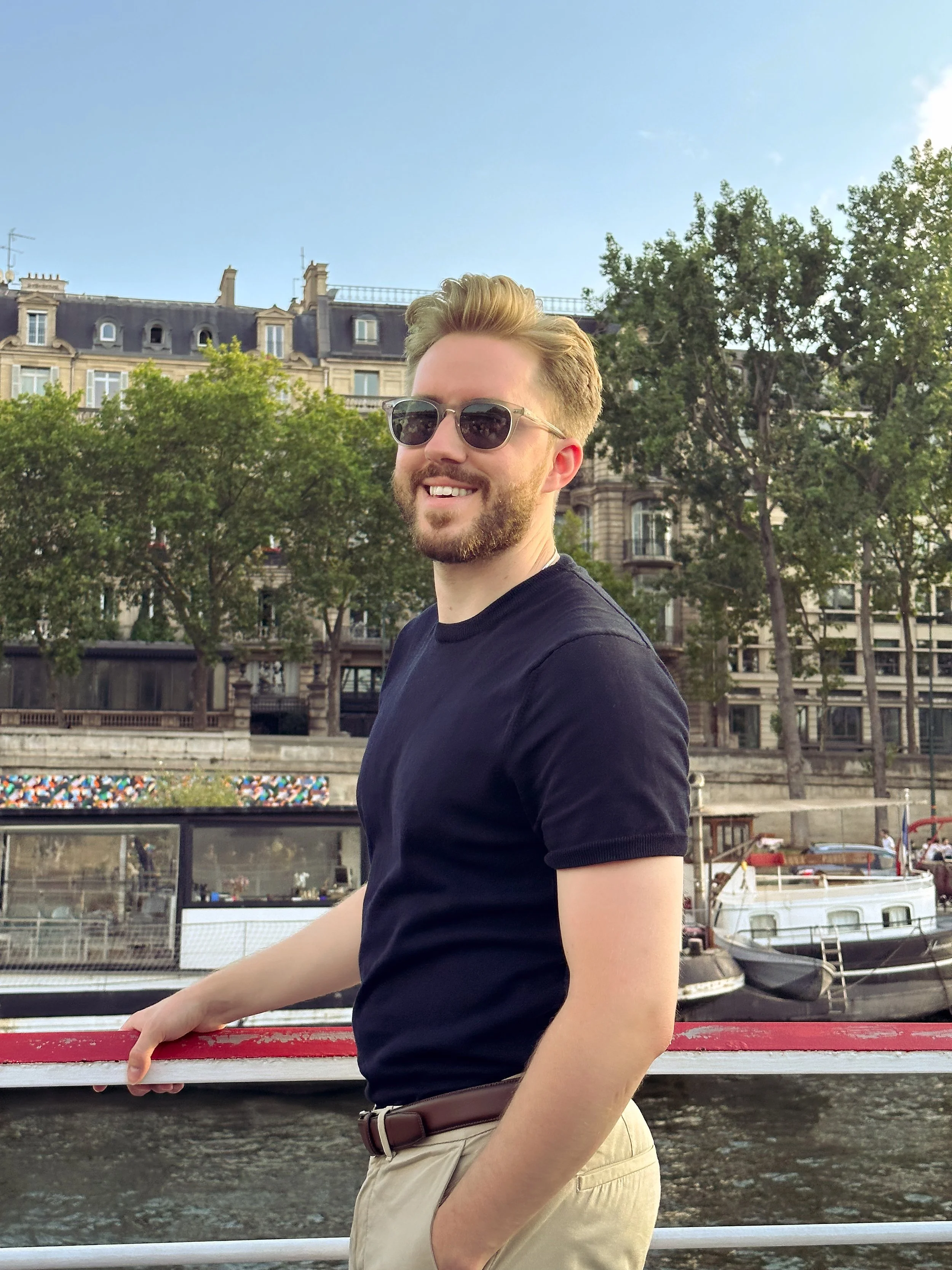 A young man with sunglasses, a beard, and styled hair, smiling while standing near a river with boats, trees, and Parisian buildings in the background during daytime.
