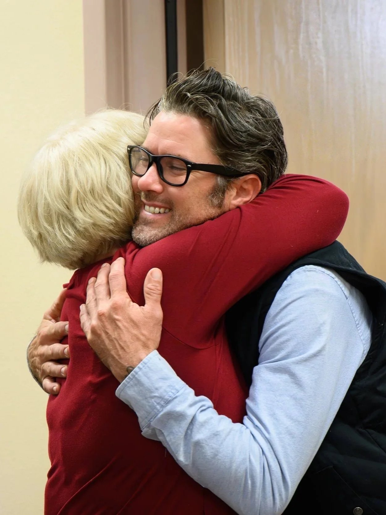 A man with dark hair and glasses hugging an older woman with blonde hair, both smiling warmly.