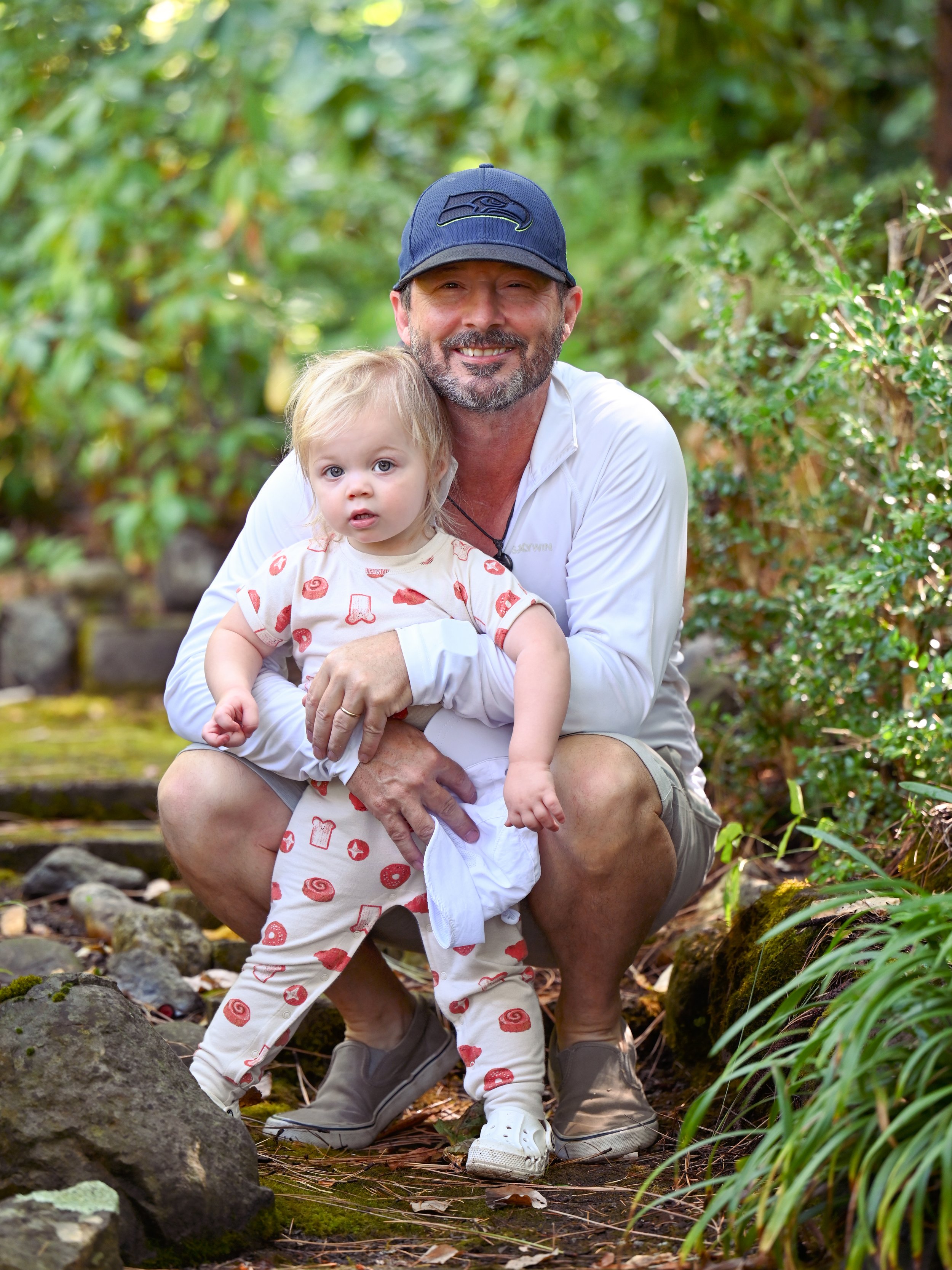 A man kneeling outdoors with a young girl, both looking at the camera, surrounded by green foliage.