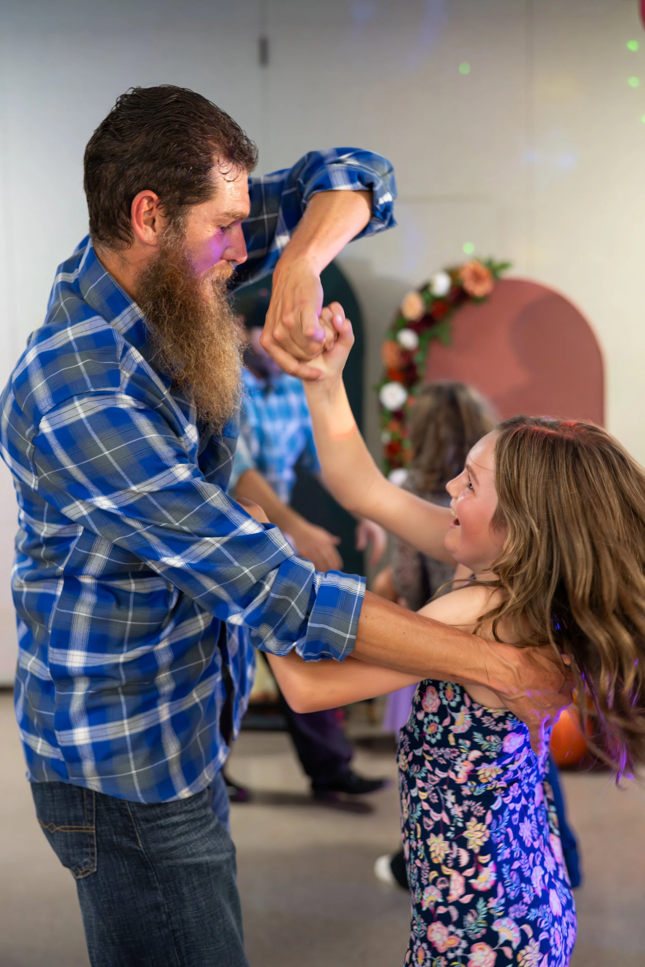 A man with a beard and long hair dancing with a young girl in a floral dress at a festive gathering. They are holding hands and smiling, with other people dancing in the background and a decorated arch in the setting.