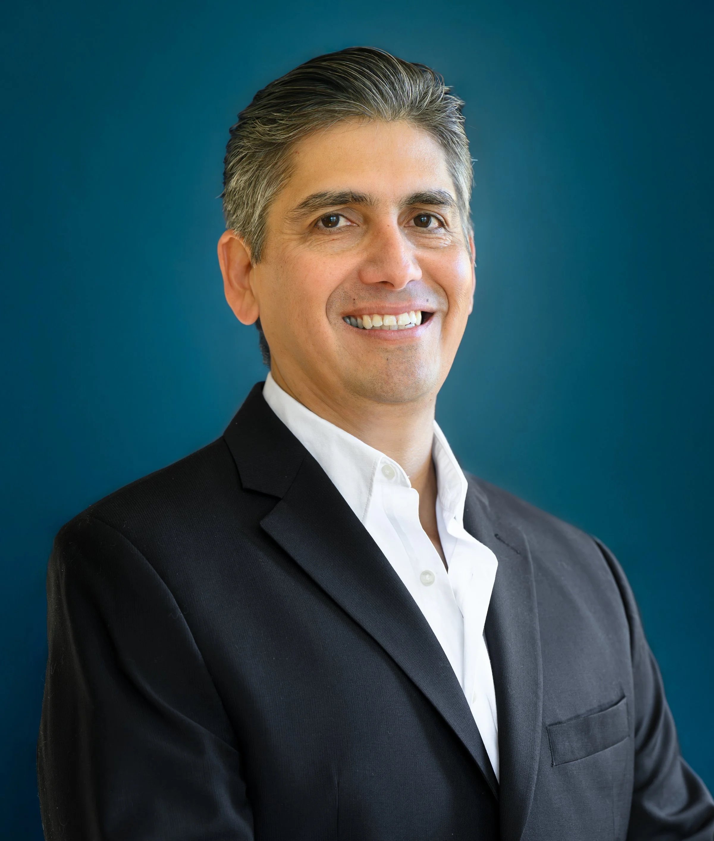 A professional man with gray hair wearing a black suit and white shirt, smiling against a blue background.