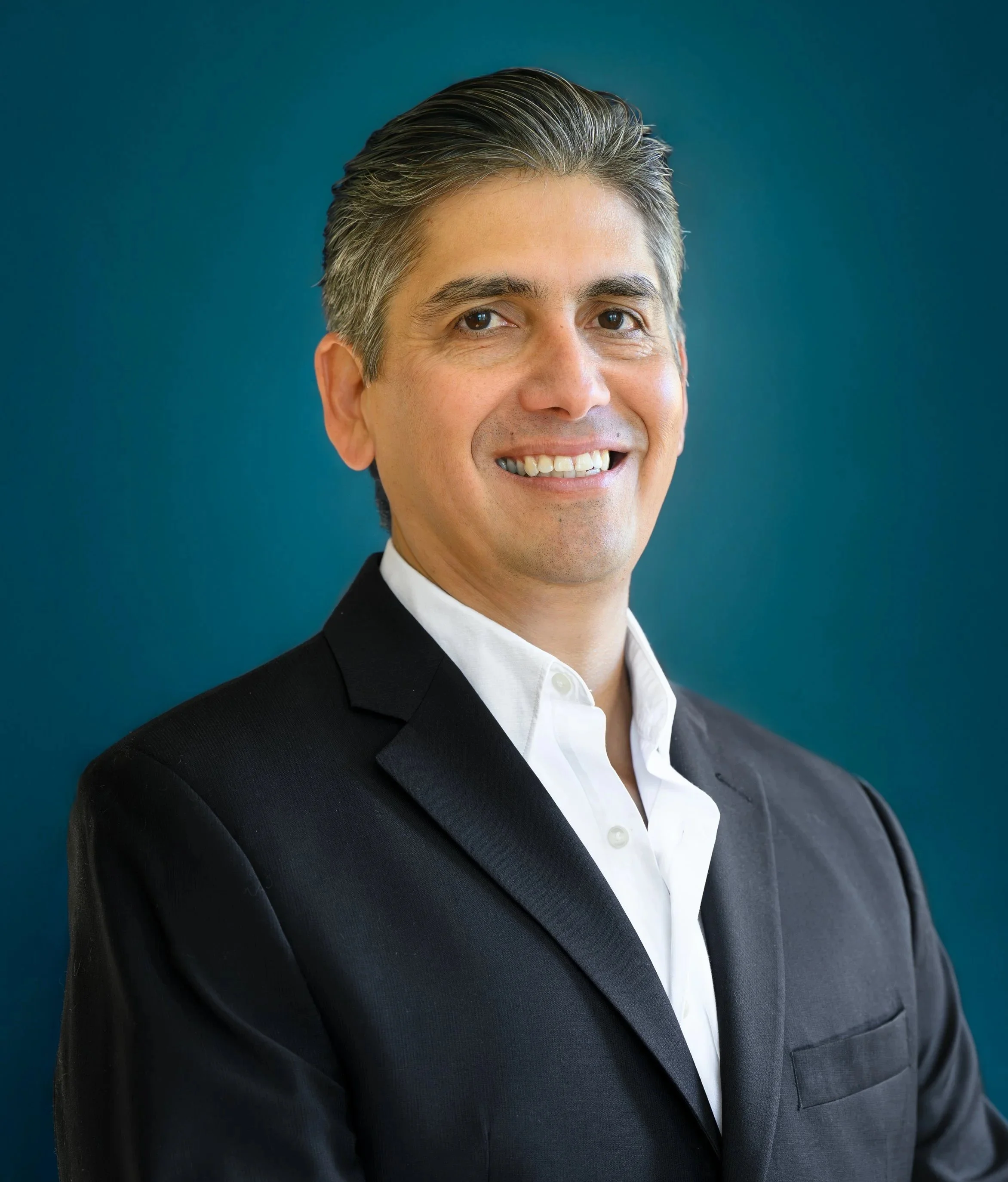 A professional man with gray hair wearing a black suit and white shirt, smiling against a blue background.