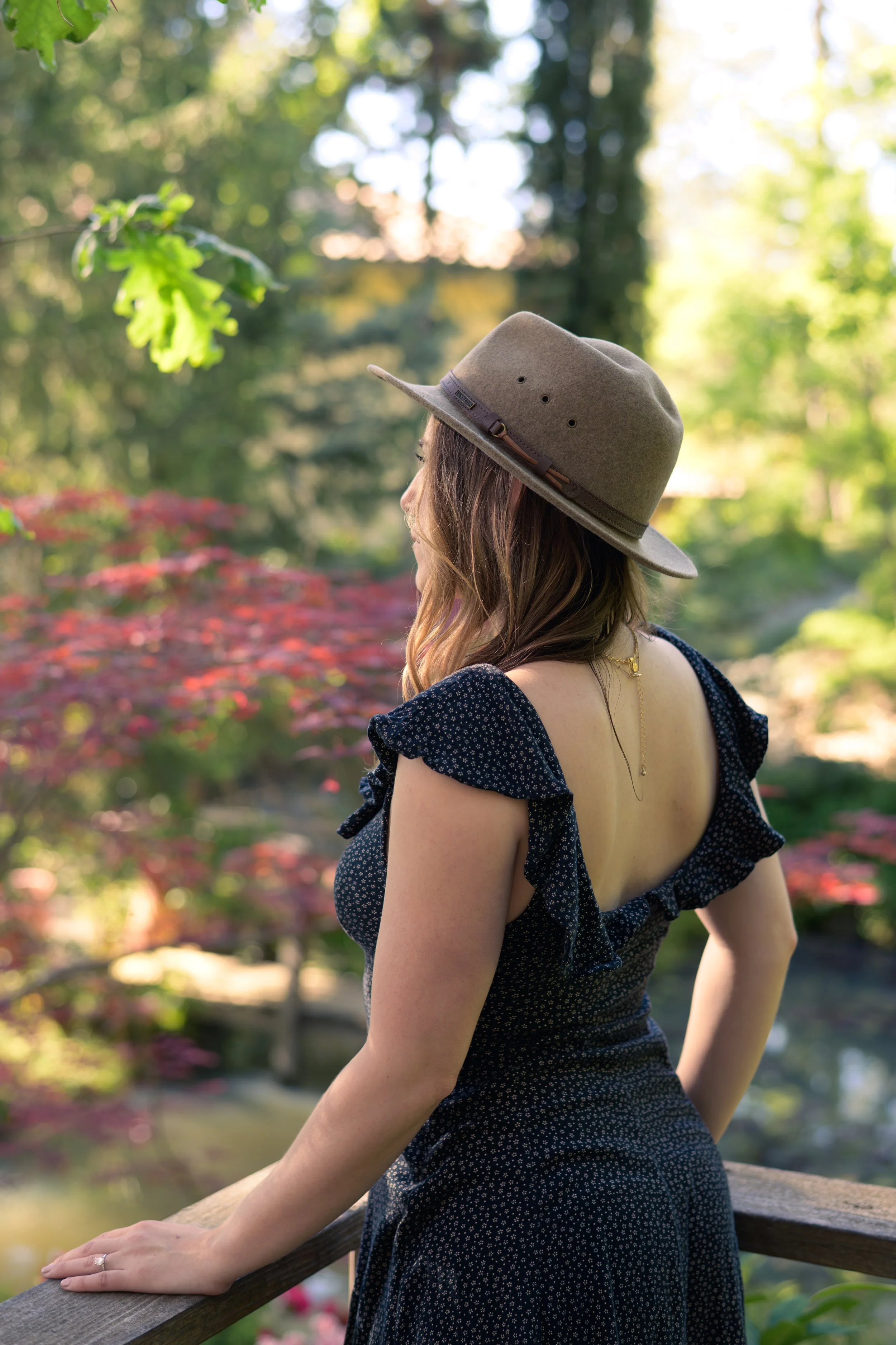 A woman with wavy brown hair in a navy dress with ruffled sleeves and a low back, wearing a gray felt hat with a brown band, stands outdoors on a wooden bridge or railing, overlooking a garden with green trees and pink flowering plants, basking in soft sunlight.