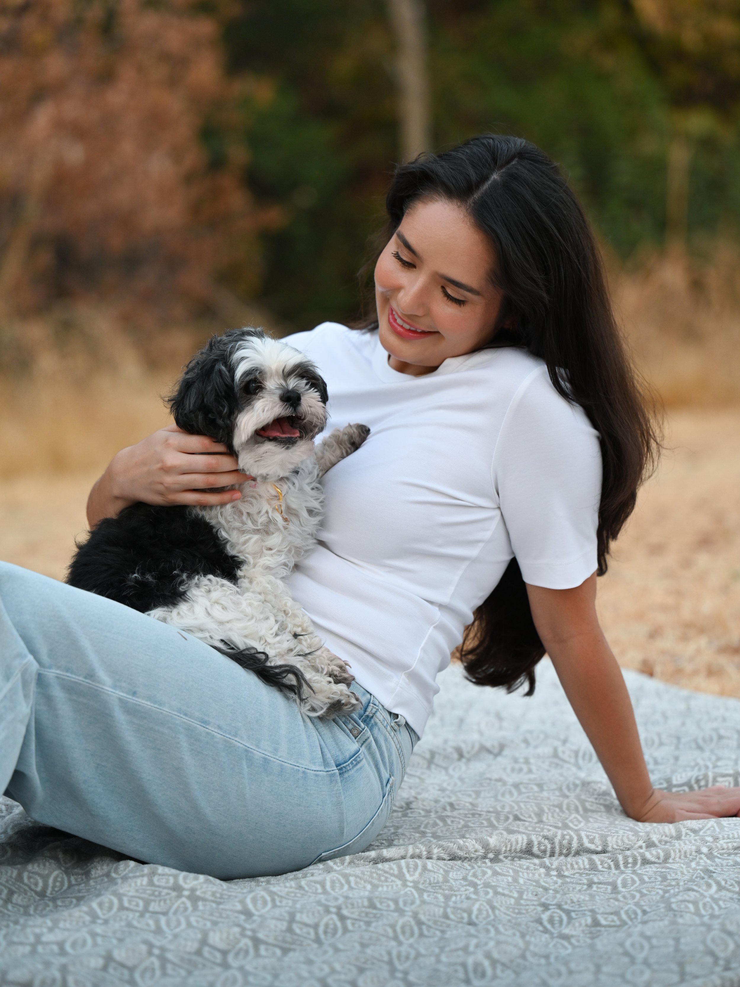 A woman sitting on a blanket outdoors, holding a small black and white dog, smiling and looking down at the dog.