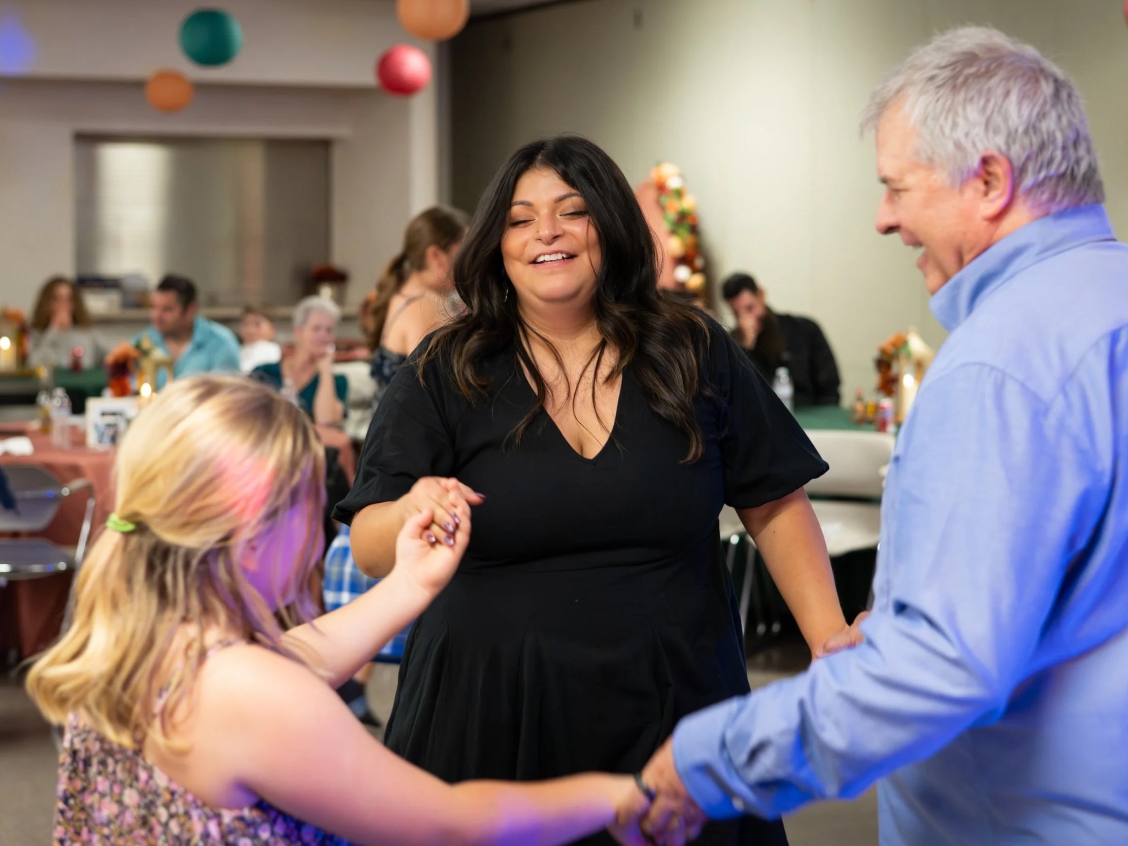 A woman in a black dress is smiling and holding hands with a girl and a man at a party or celebration. The girl has blonde hair and is wearing a floral dress, and the man is wearing a light blue shirt. There are people sitting at tables in the background with festive decorations.
