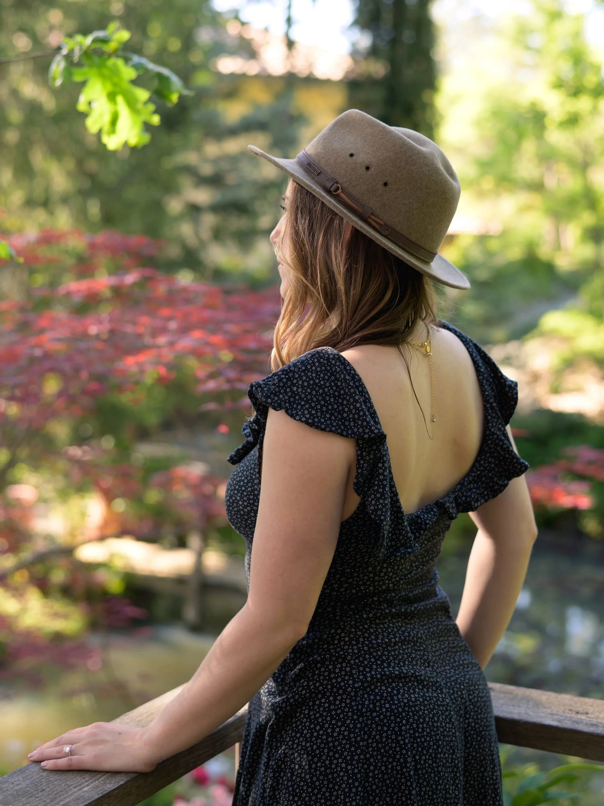 A woman with wavy brown hair wearing a wide-brimmed beige hat and a black dress with small white polka dots, standing outdoors on a wooden railing in a lush, green park with trees and red flowering plants.