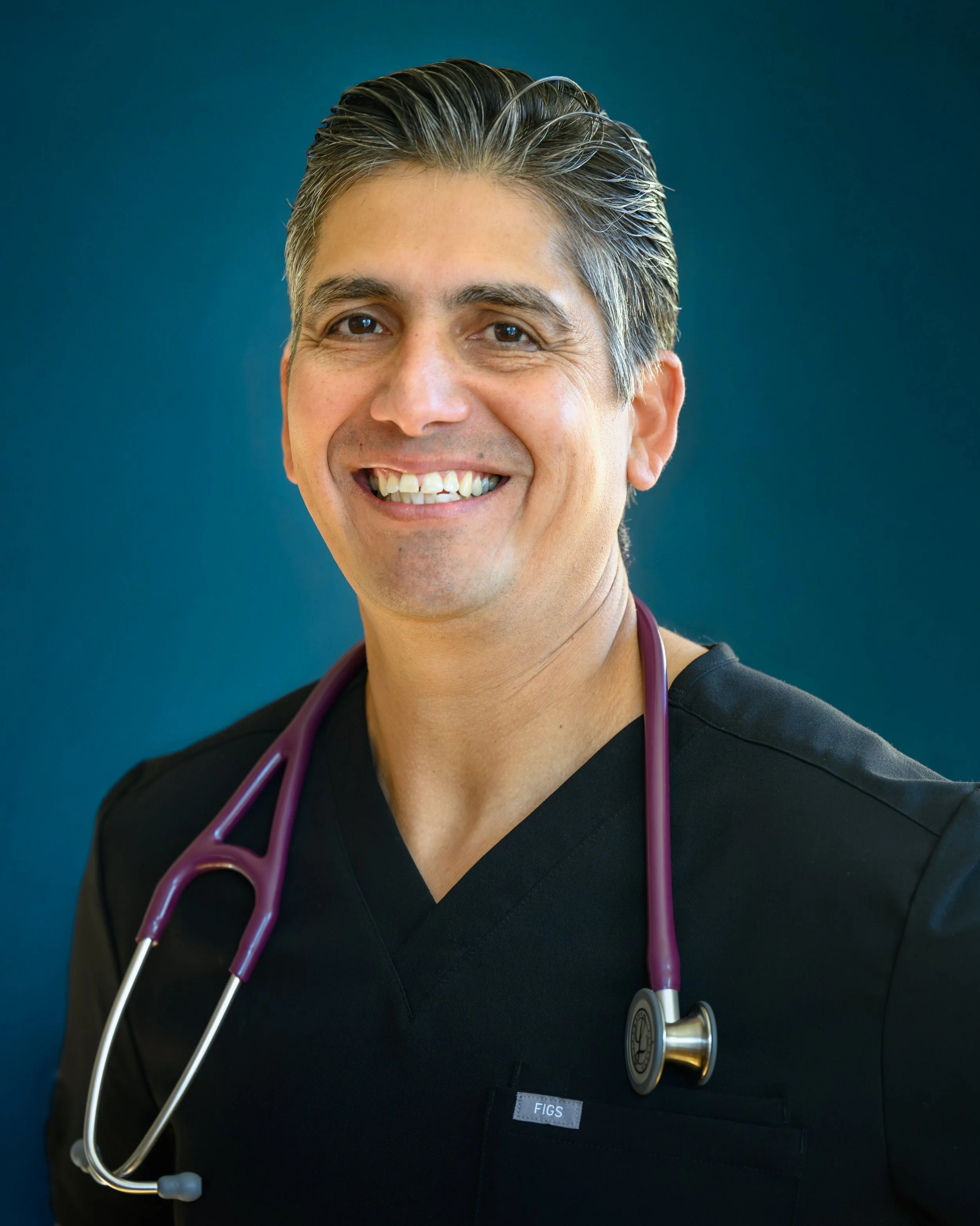 A smiling male healthcare professional wearing black scrubs and a purple stethoscope around his neck.