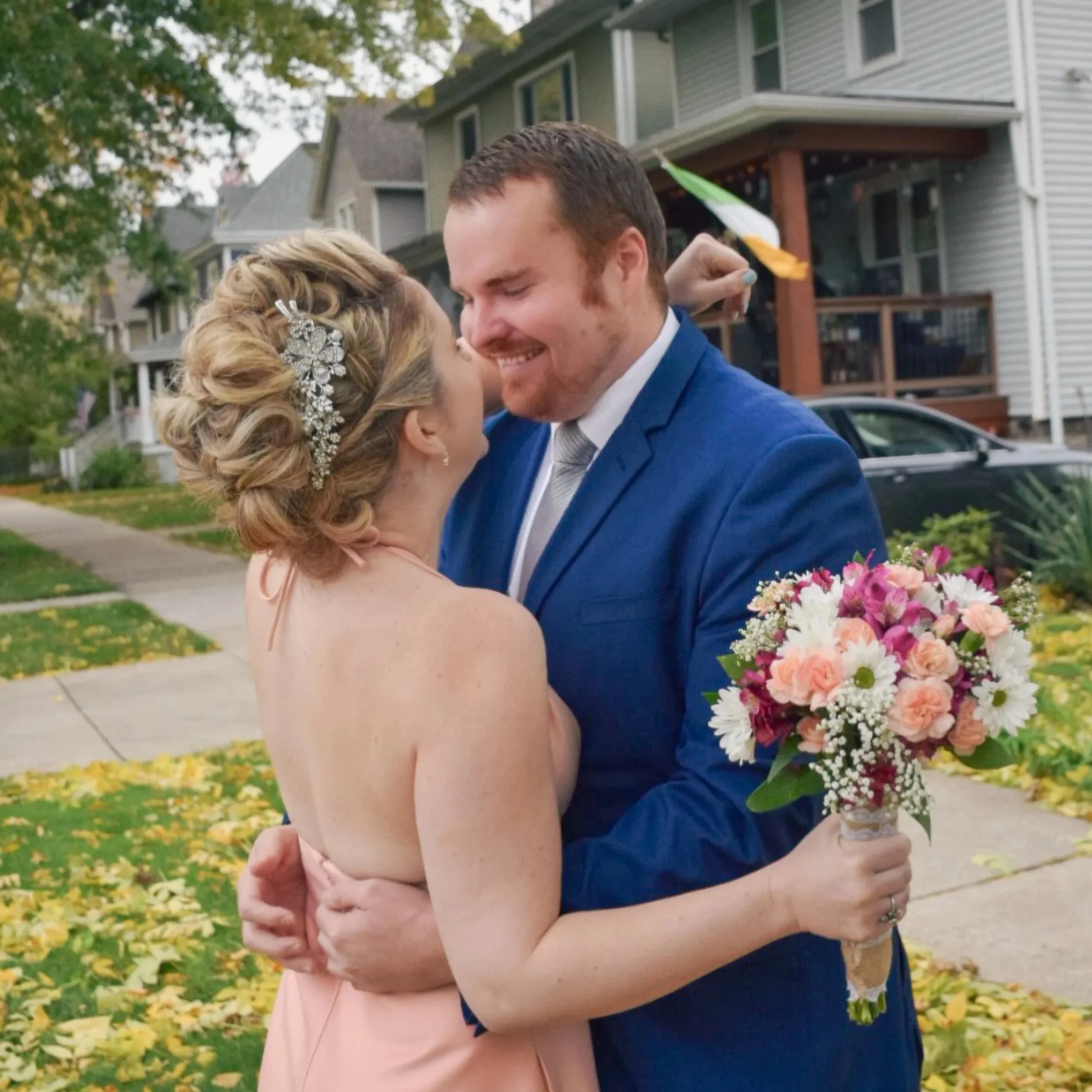 A newlywed couple happily embracing outdoors in a neighborhood with houses and autumn leaves on the ground. The bride holds a bouquet of pink and white flowers, and the groom wears a blue suit.