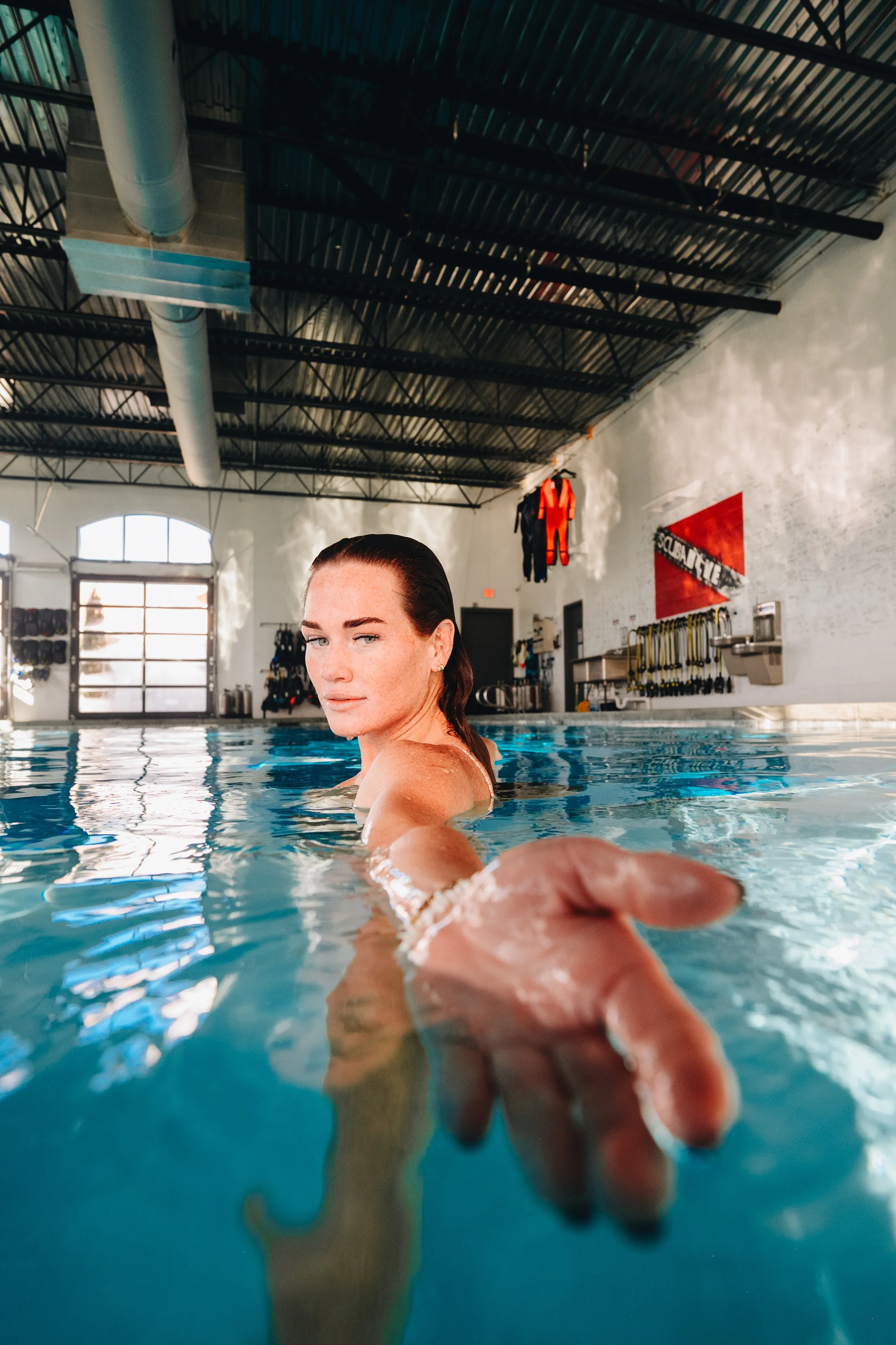 A woman in a swimming pool reaching out towards the camera with one hand, inside an indoor swimming pool facility.