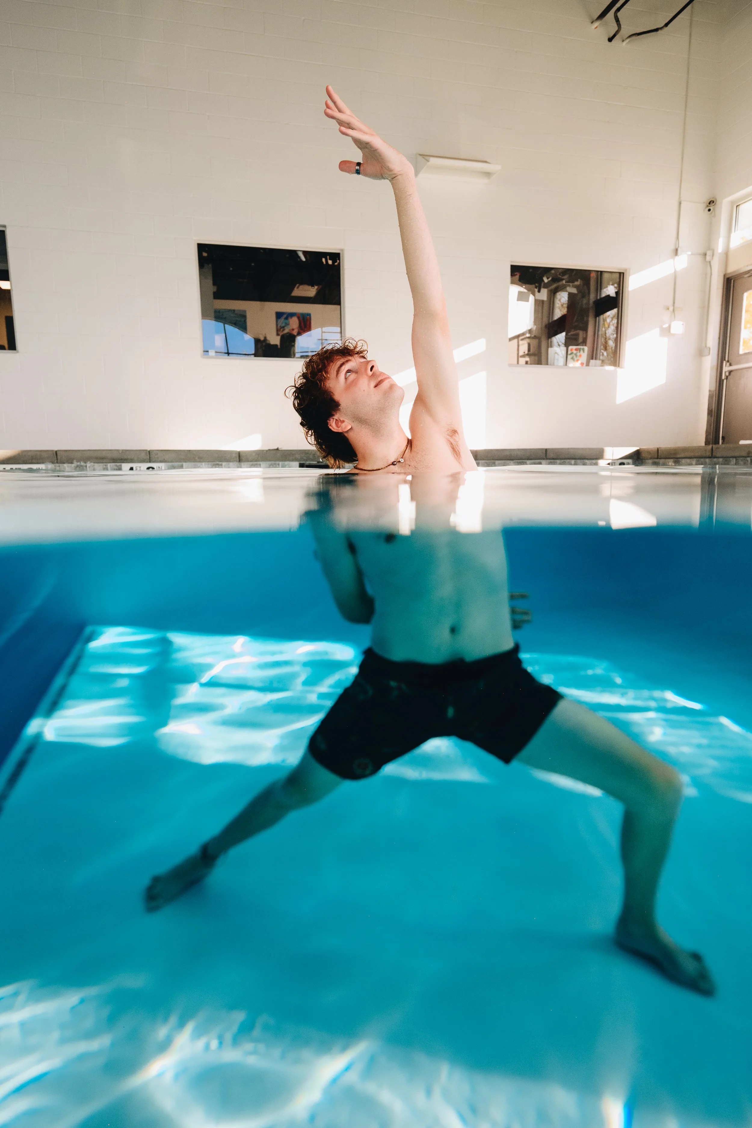 A young man in black swim trunks kneels underwater in a swimming pool, with one arm reaching up above the water's surface and the other arm extended behind him. The indoor pool area has a white brick wall with windows and sunlight streaming in.