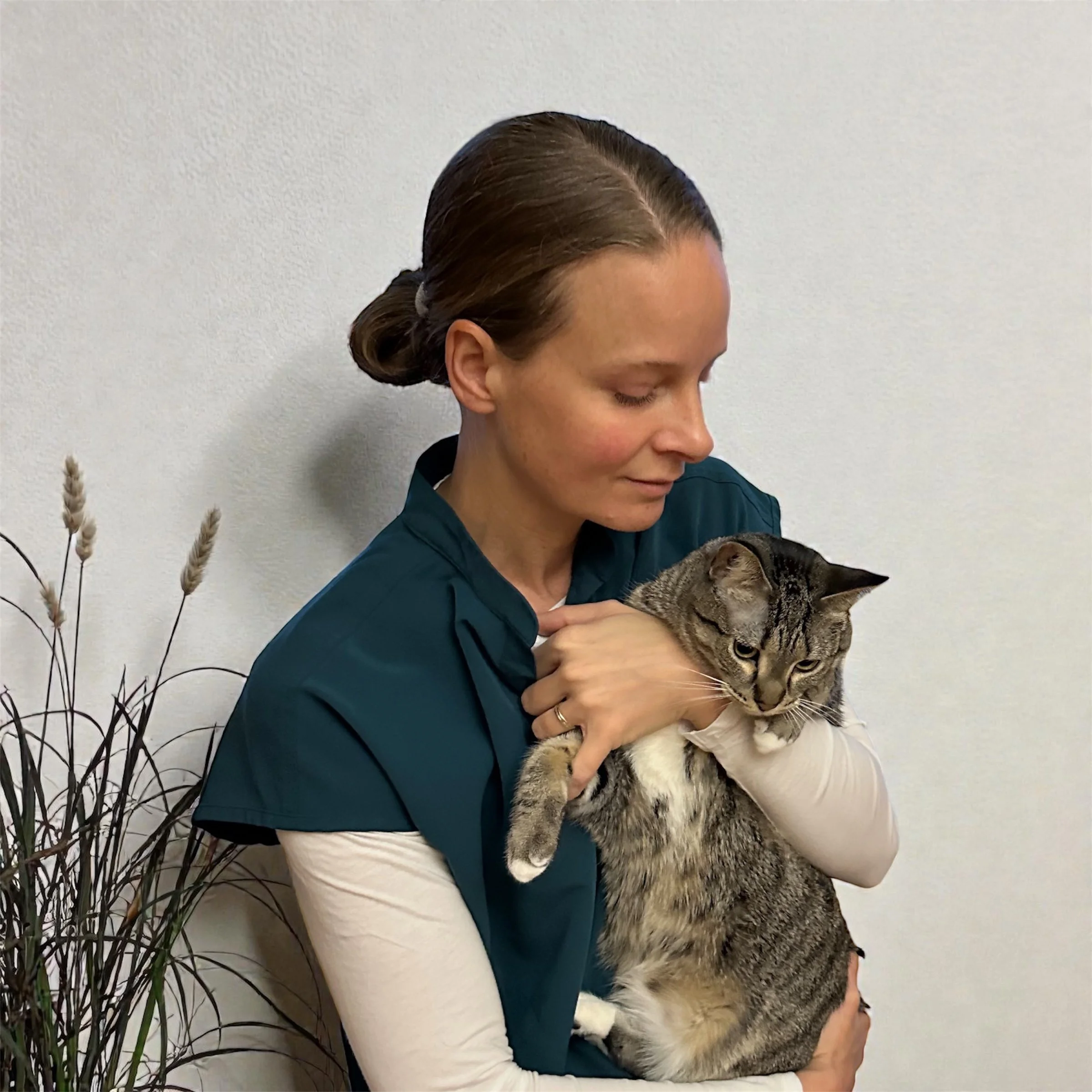 A woman with brown hair in a bun wearing a teal medical uniform holding a tabby cat with a white chest against a plain white wall.