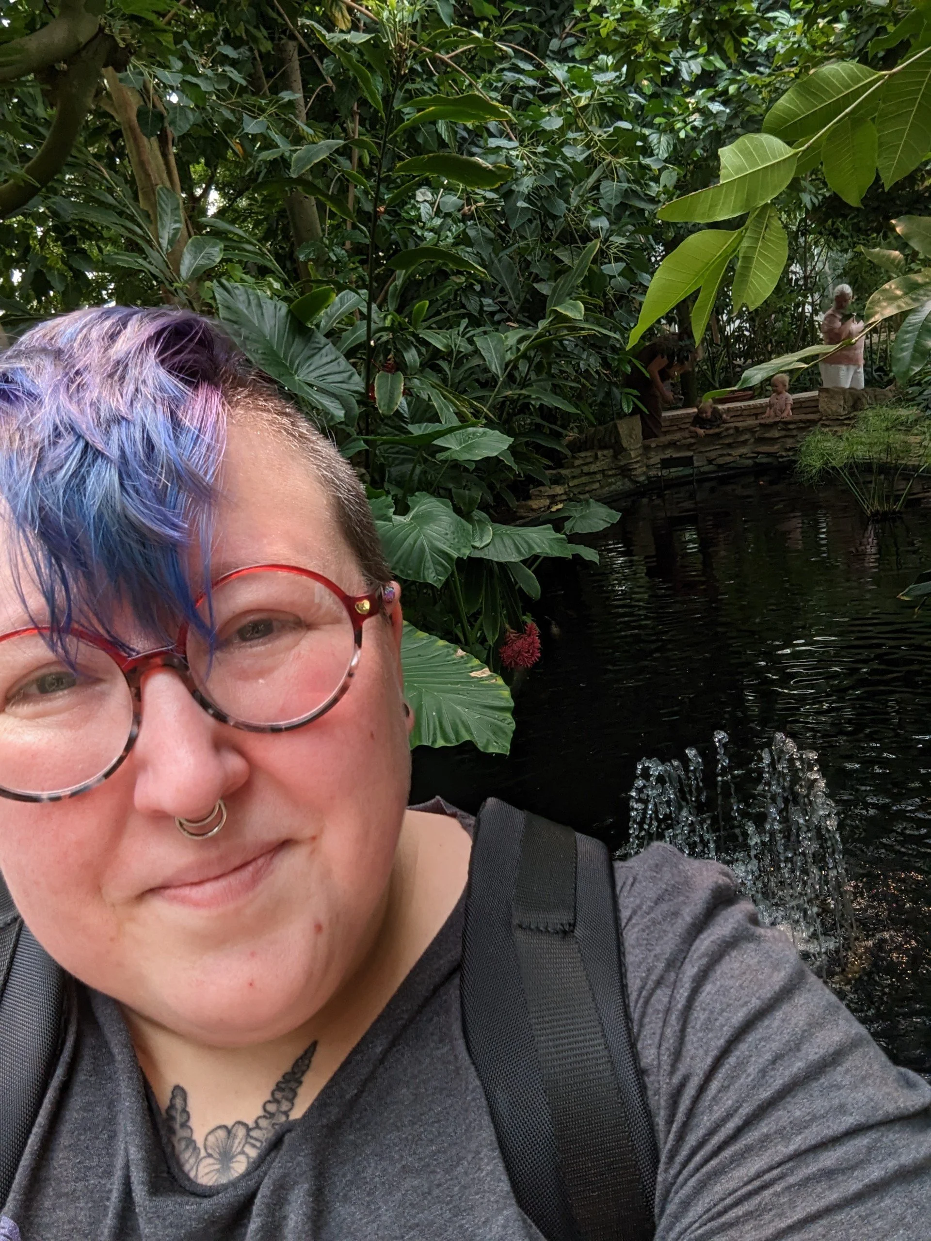A person with colorful hair and glasses taking a selfie near a pond in a lush garden with a small stone bridge and some people in the background.