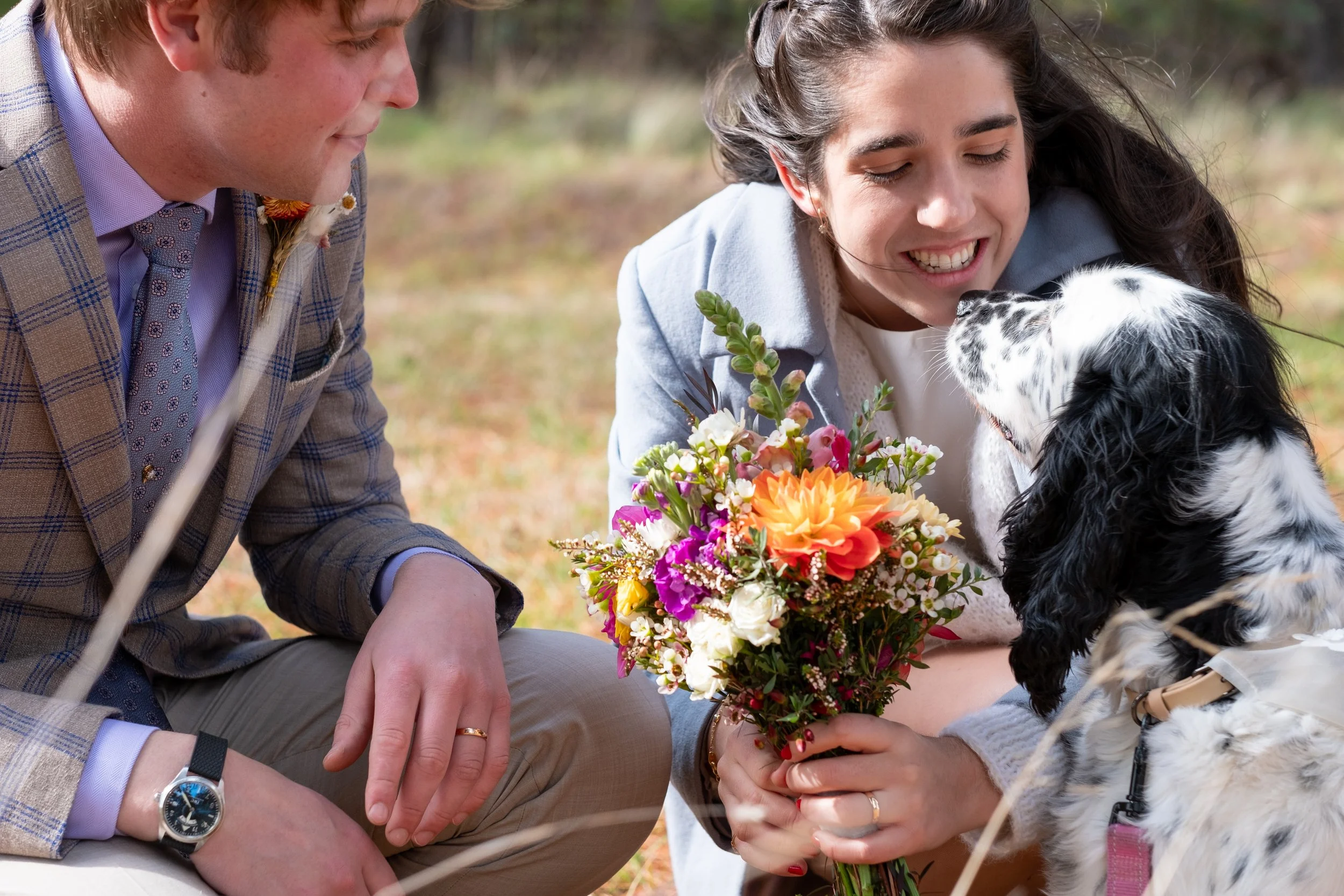 A woman holding a colorful bouquet of flowers, smiling at her dog. A man is sitting nearby, looking on. The scene is outdoors with grass and trees in the background.